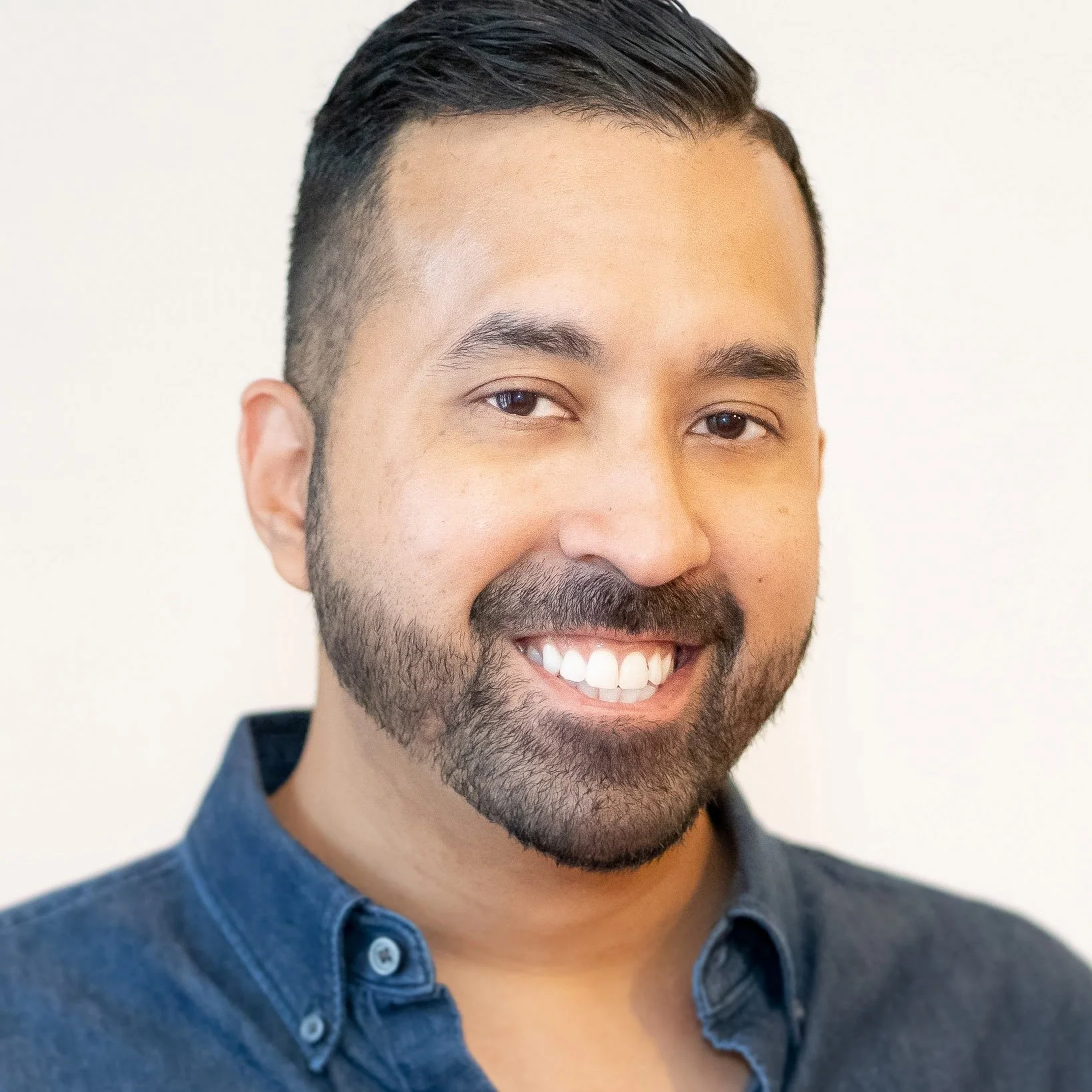 Close-up portrait of a smiling man with short dark hair, facial hair, and wearing a denim shirt against a plain background.