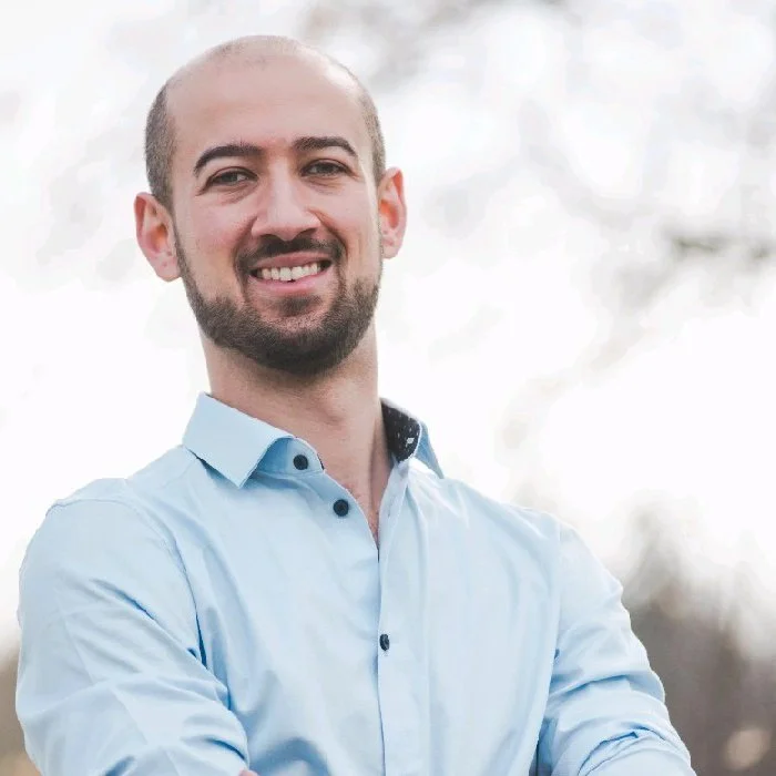 A smiling man with a beard and short hair, wearing a light blue button-up shirt, standing outdoors with blurred trees in the background.