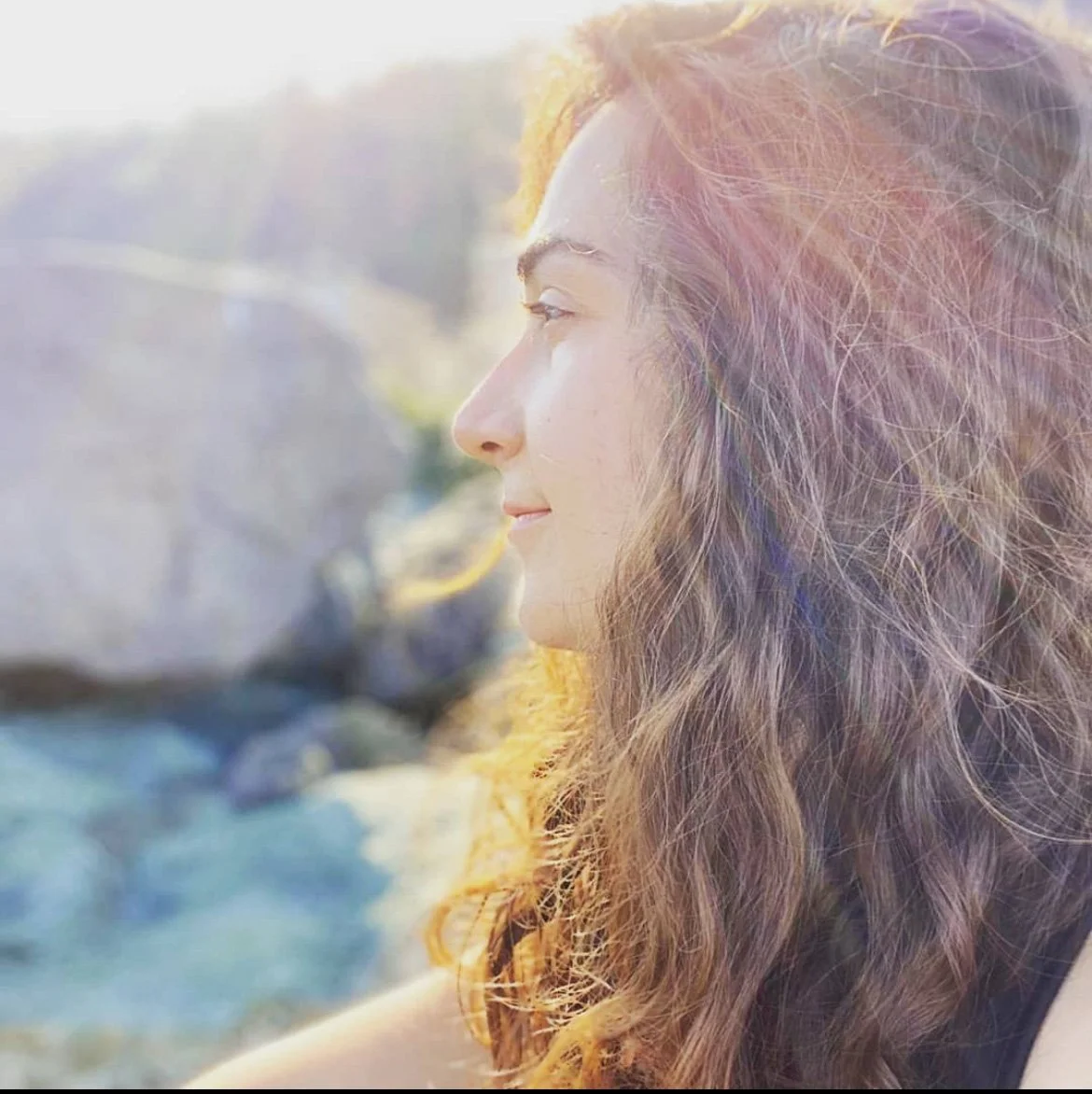A woman with wavy hair looking towards the sun, with a blurred background of rocks and water.