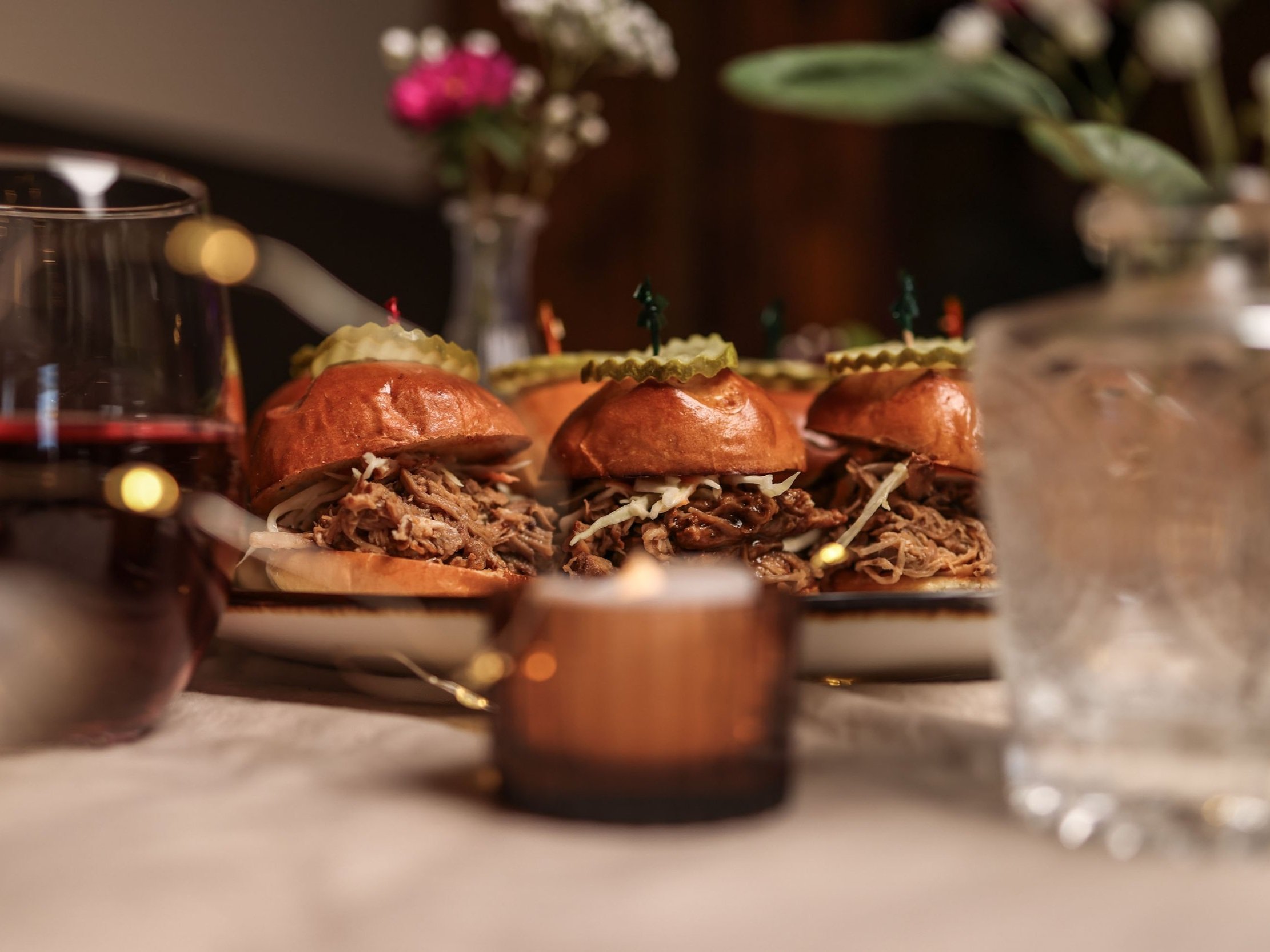  Close-up of pulled pork sandwiches on a table adorned with candles and glasses of red wine, creating an intimate dining atmosphere. 