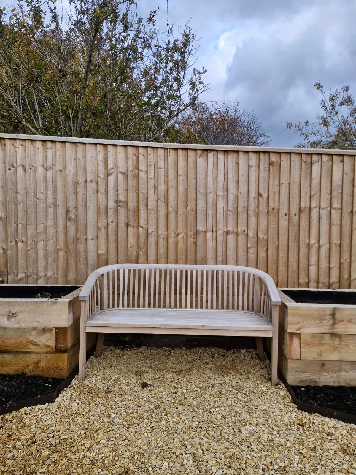 A backyard scene with a wooden privacy fence, a white wooden bench, and two large wooden planter boxes with soil, set on a gravel surface under a cloudy sky.