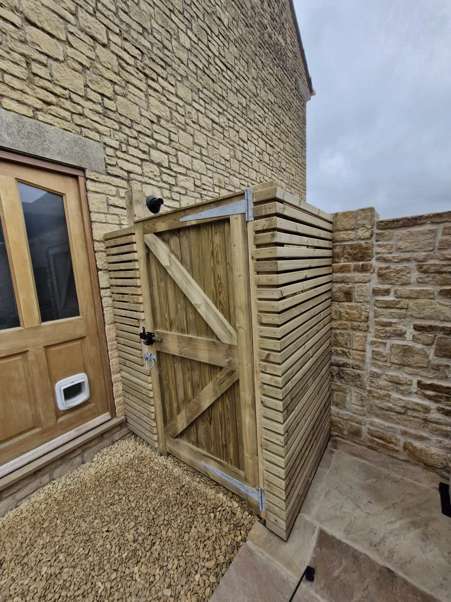 A wooden outdoor structure next to a brick wall and a wooden door, with gravel on the ground and a cloudy sky above.