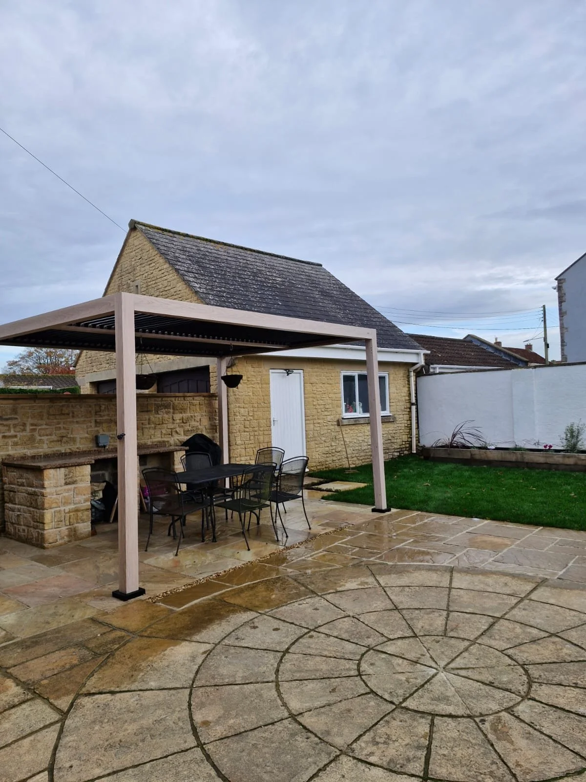 A backyard patio with a circular stone design, a small grassy area, a beige brick house with a white door and window, and a wooden pergola with outdoor furniture underneath during daytime with cloudy sky.