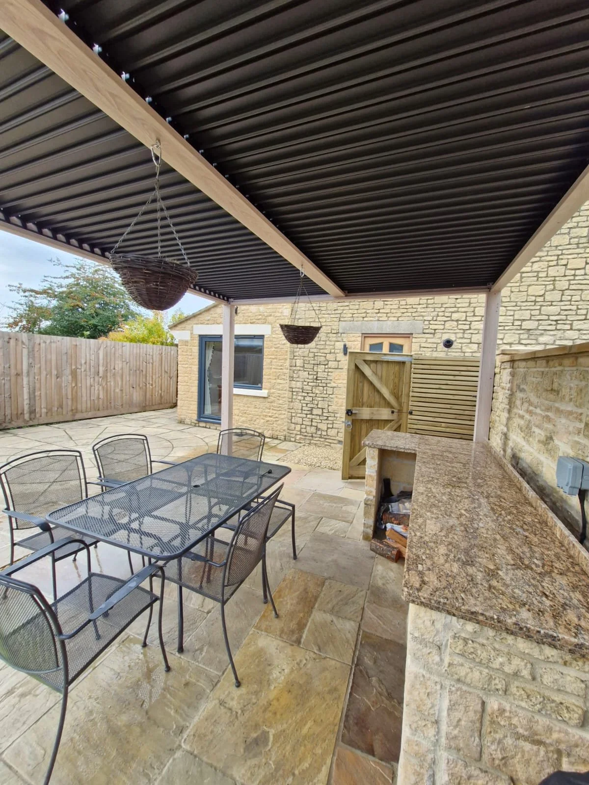 Outdoor patio area with a metal dining table and six chairs, wicker hanging planters, a granite countertop bar, a stone building with a window, a wooden gate, a brick wall, and cloudy sky.