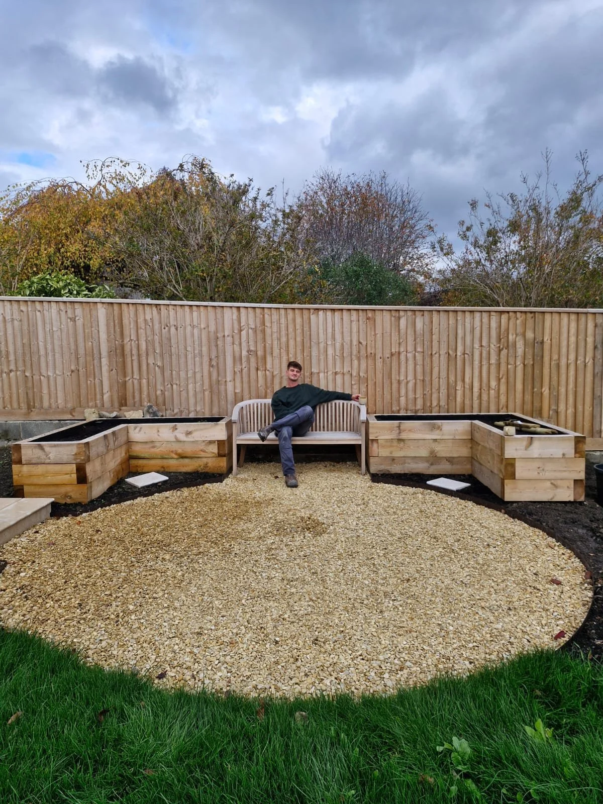 A person sitting on a wooden garden bench in a backyard with raised garden beds, gravel ground cover, a wooden fence, and trees with fall foliage under a cloudy sky.