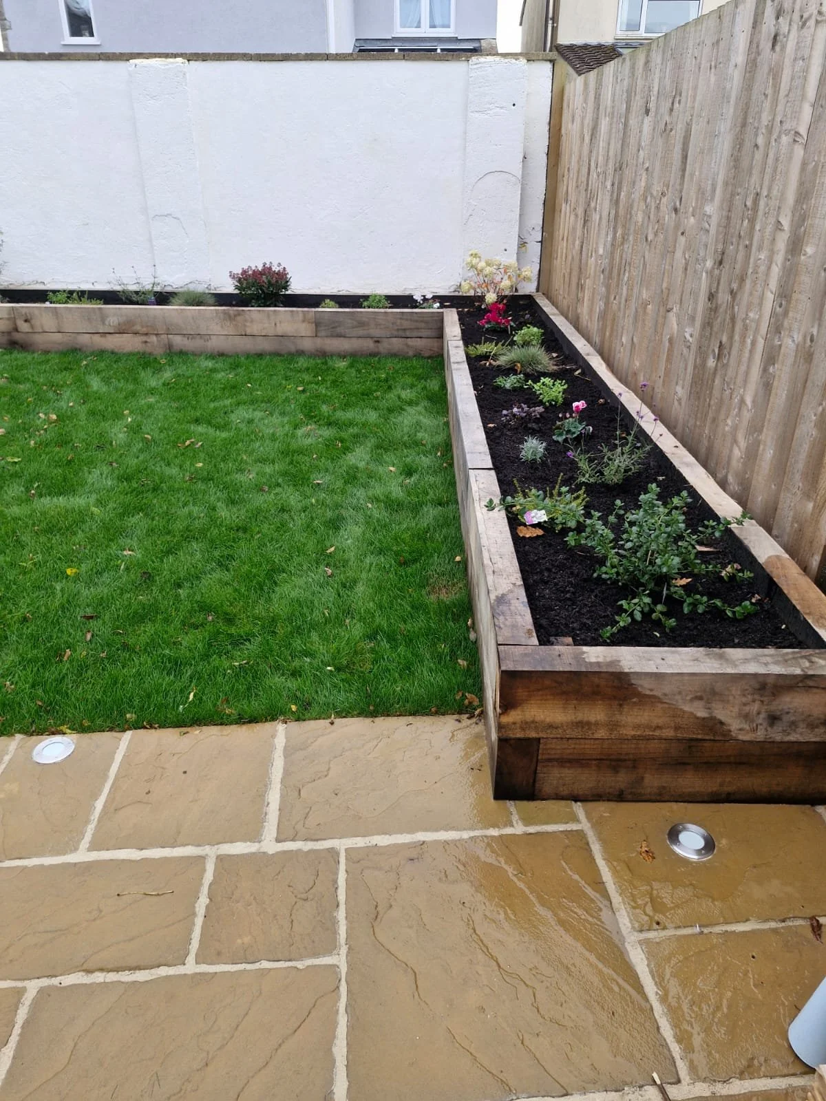 A small garden with a wooden raised flower bed filled with various plants and flowers, on a patio with beige stone tiles and a green lawn. A white wall and a wooden fence are in the background.