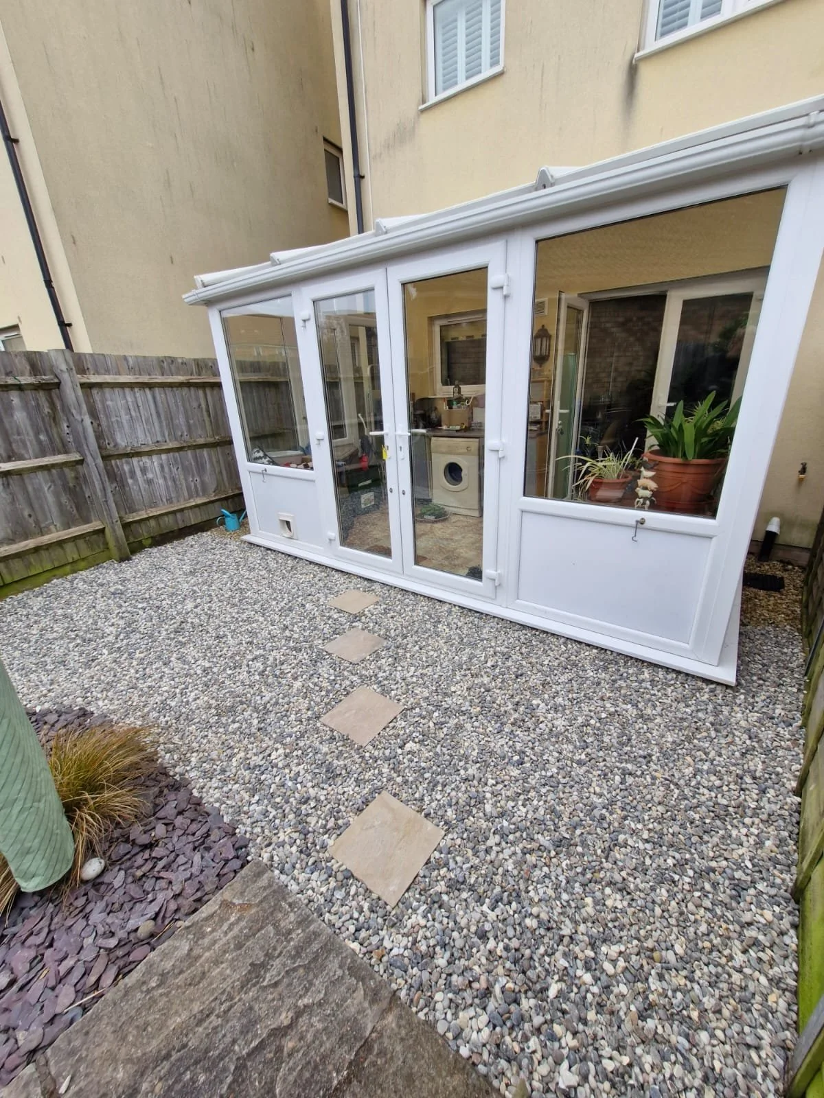 A small backyard with a gravel surface and a wooden fence. There is a white conservatory attached to a beige house, with potted plants visible inside. Several stepping stones lead across the gravel. Part of a cactus is visible in the lower left corne