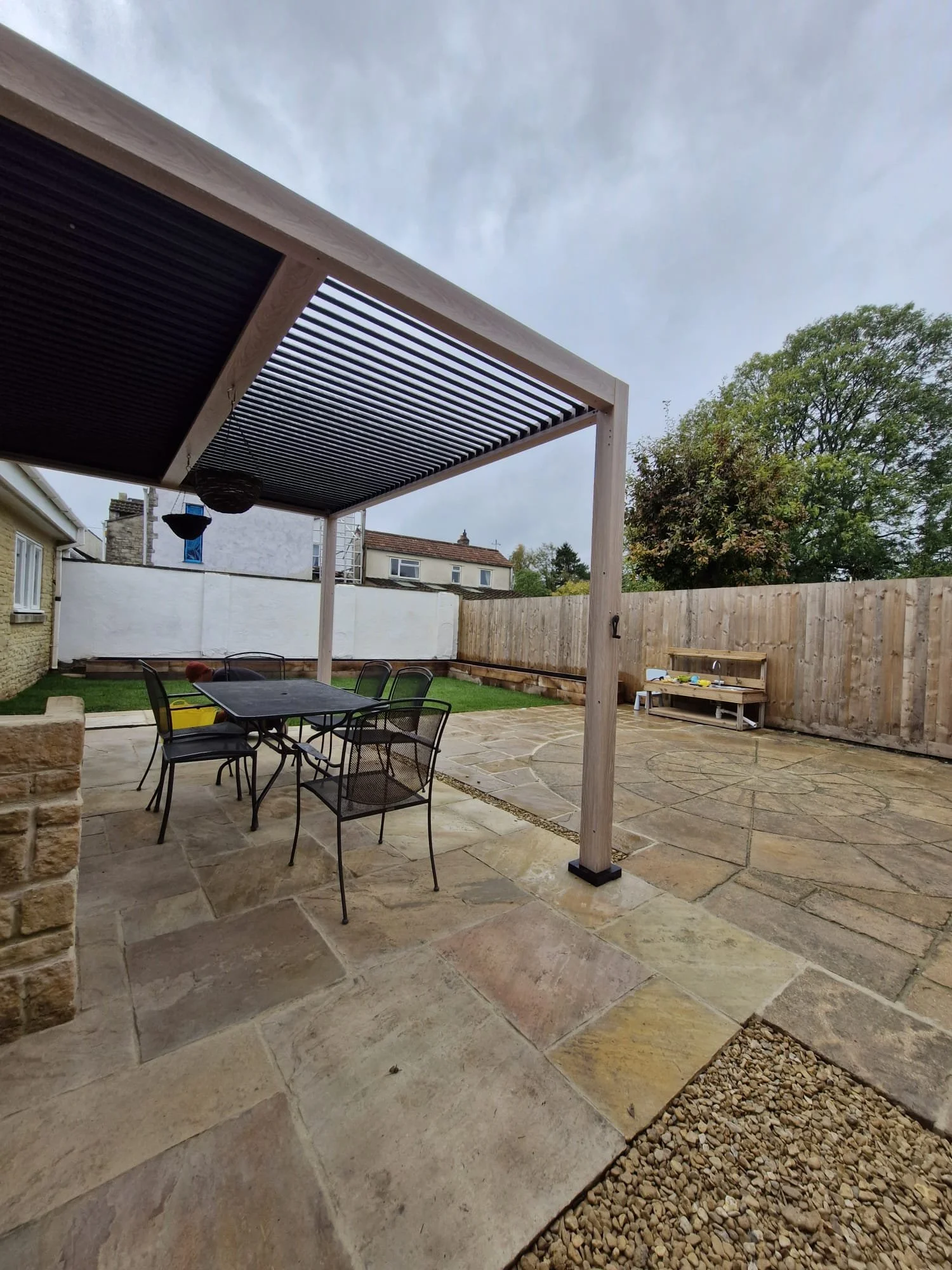 Backyard patio with stone paving, outdoor dining table with chairs, wooden pergola with slatted roof, fenced yard with trees and neighboring houses, overcast sky.