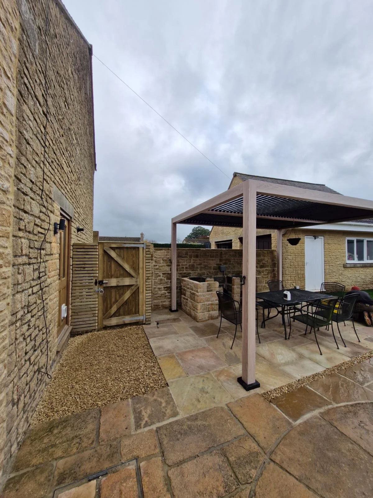 An outdoor patio area with stone paving, a dining table with six chairs under a wooden pergola, and a brick barbecue grill. A wooden gate and a wall with a window are visible in the background, and the sky is overcast.