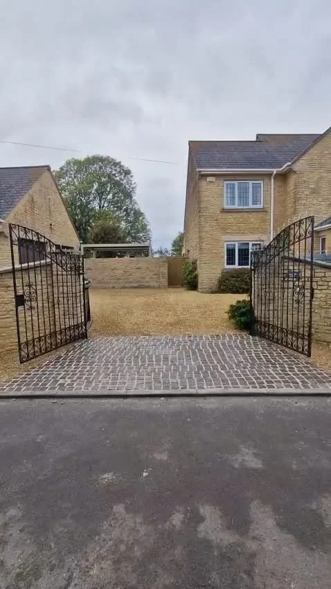 View of a brick-paved driveway leading through an ornate black gate, with a yellow brick house and a lawn in the background under a cloudy sky.