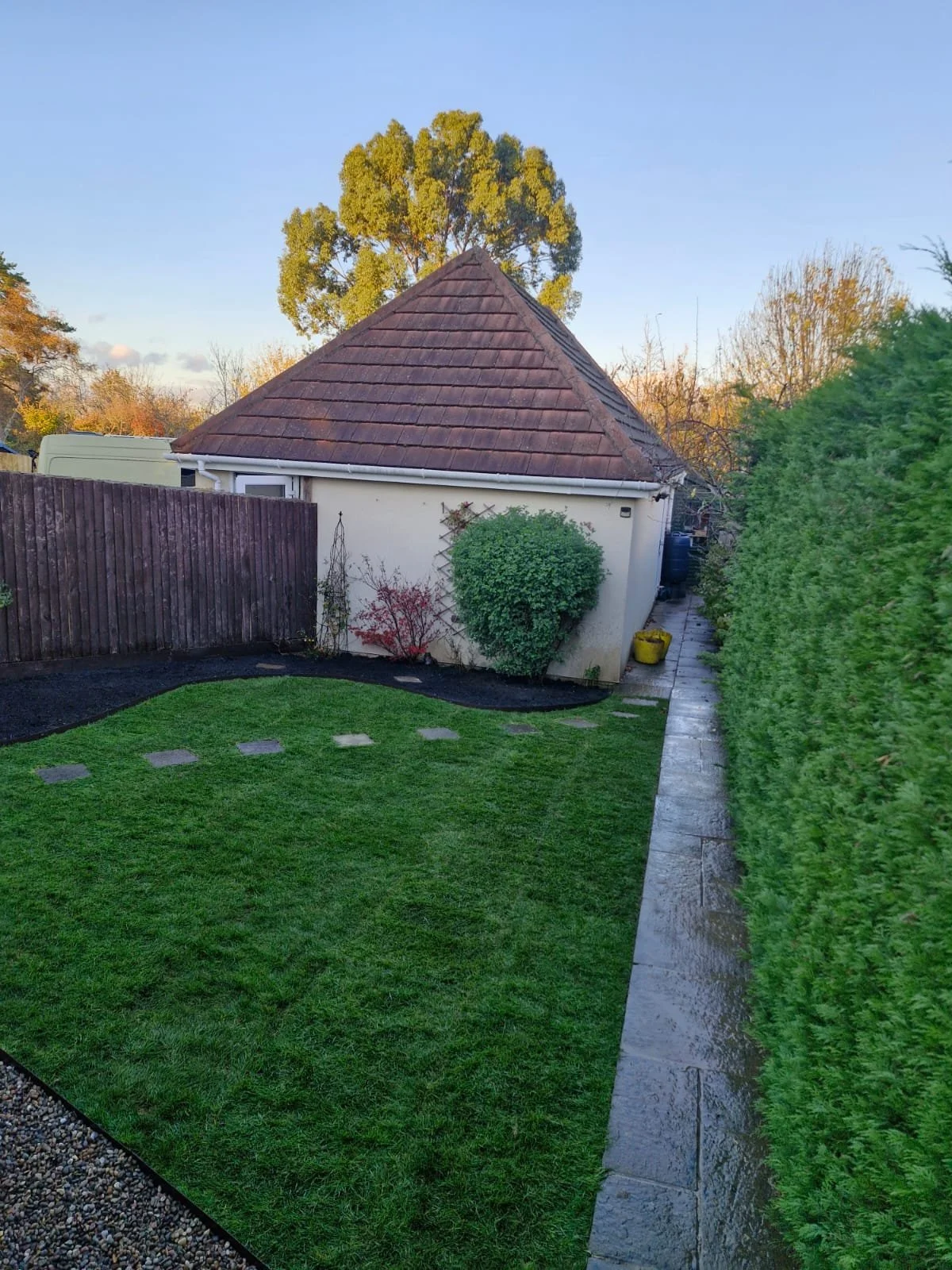 View of a backyard with a small white building with a brown tiled roof, green lawn, and hedge on the right side. There's a wooden fence on the left and a paved pathway along the hedge. Trees in the background with clear blue sky.