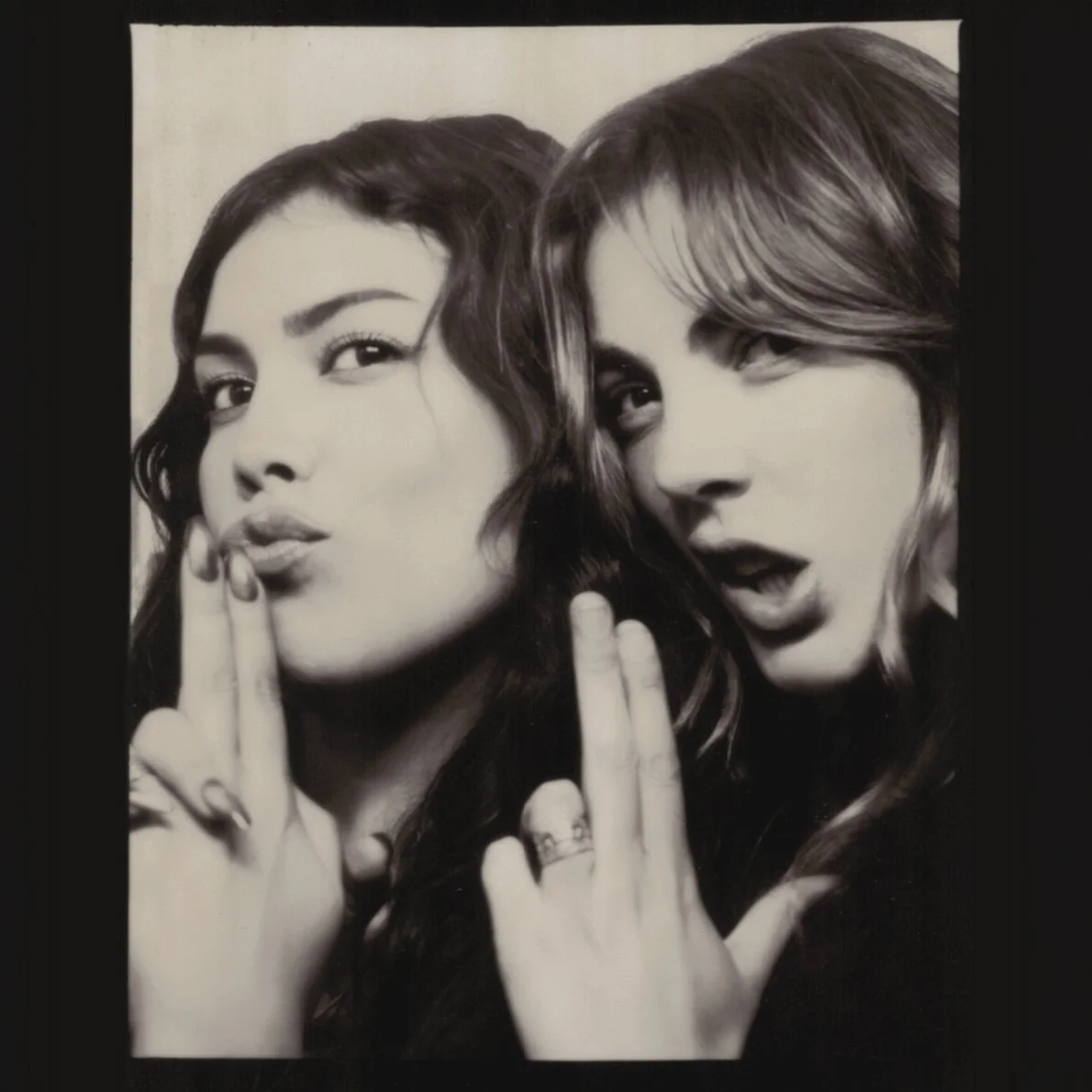 Black and white photo of two women making playful facial expressions and holding up their middle fingers. One woman has dark hair and is making a shushing gesture, while the other has light hair and has her mouth slightly open.