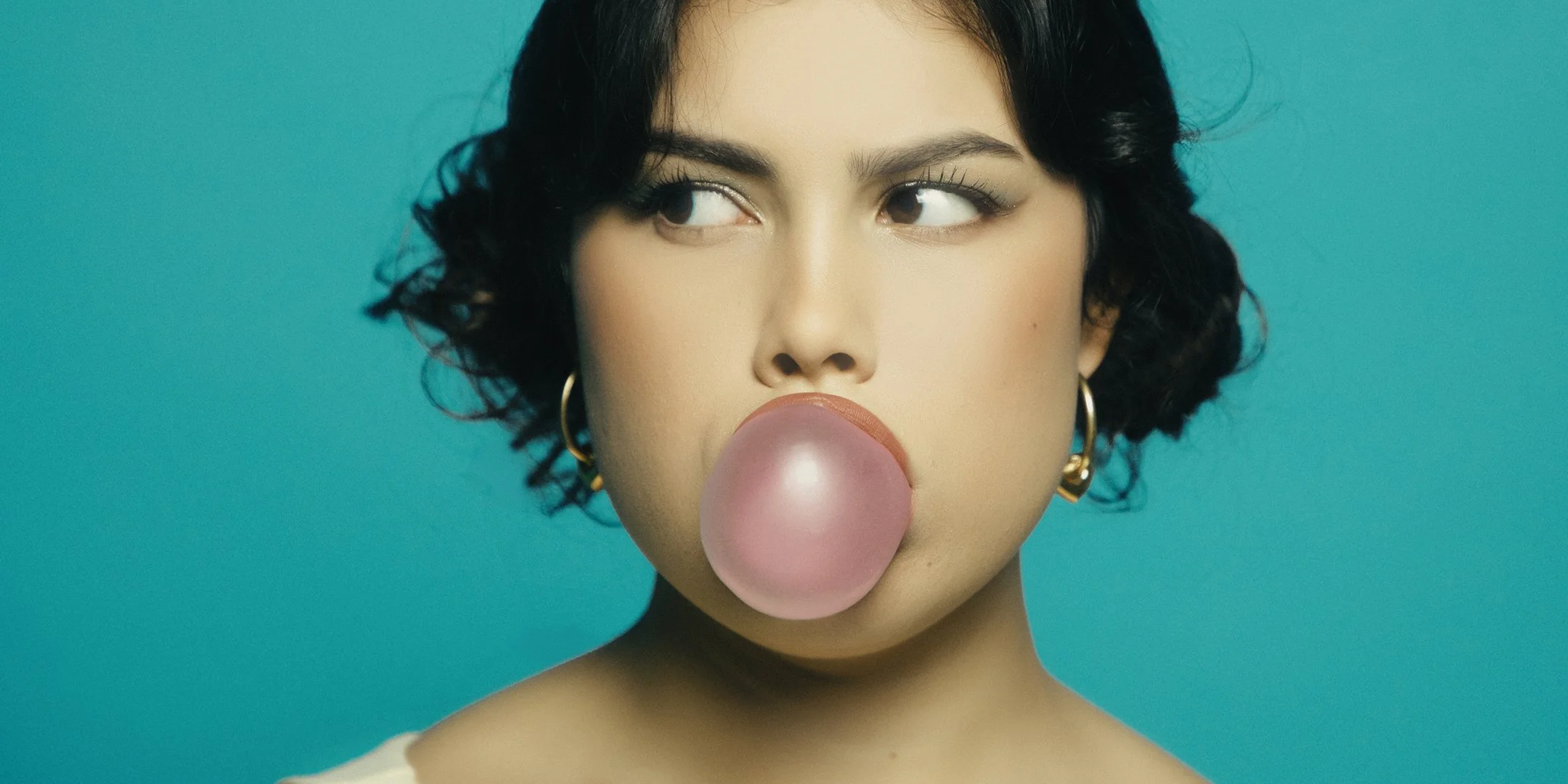 A woman with dark, curly hair blowing a pink bubblegum bubble against a teal background.
