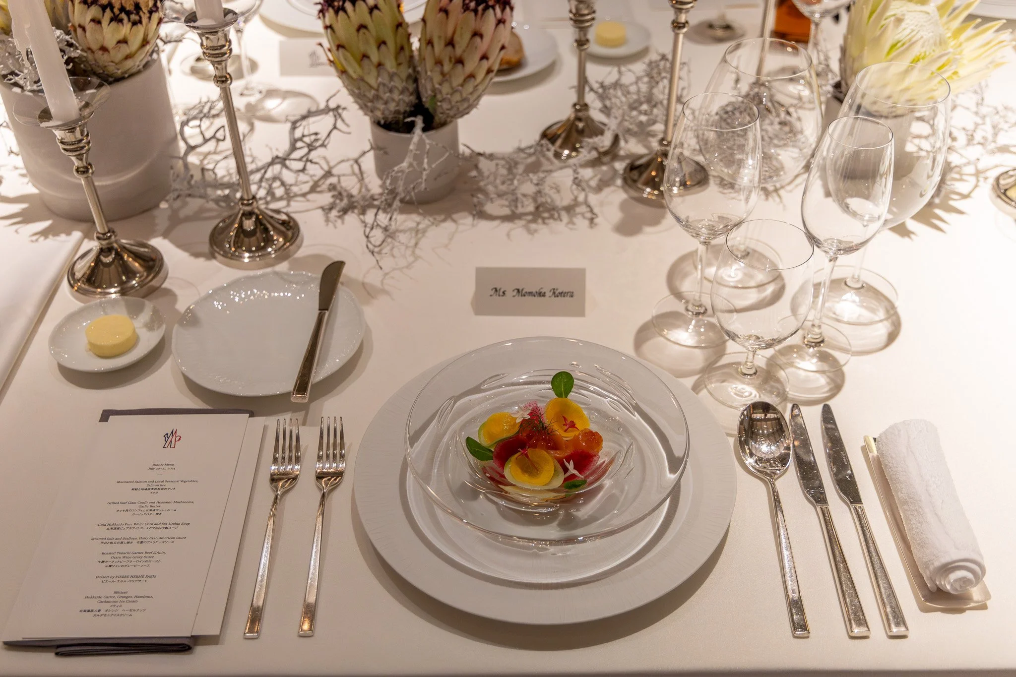 Elegant formal dinner table setting with a glass bowl of colorful edible flowers and garnish, multiple wine glasses, silverware, a menu, a rolled white napkin, candles, artichokes, and table decorations.