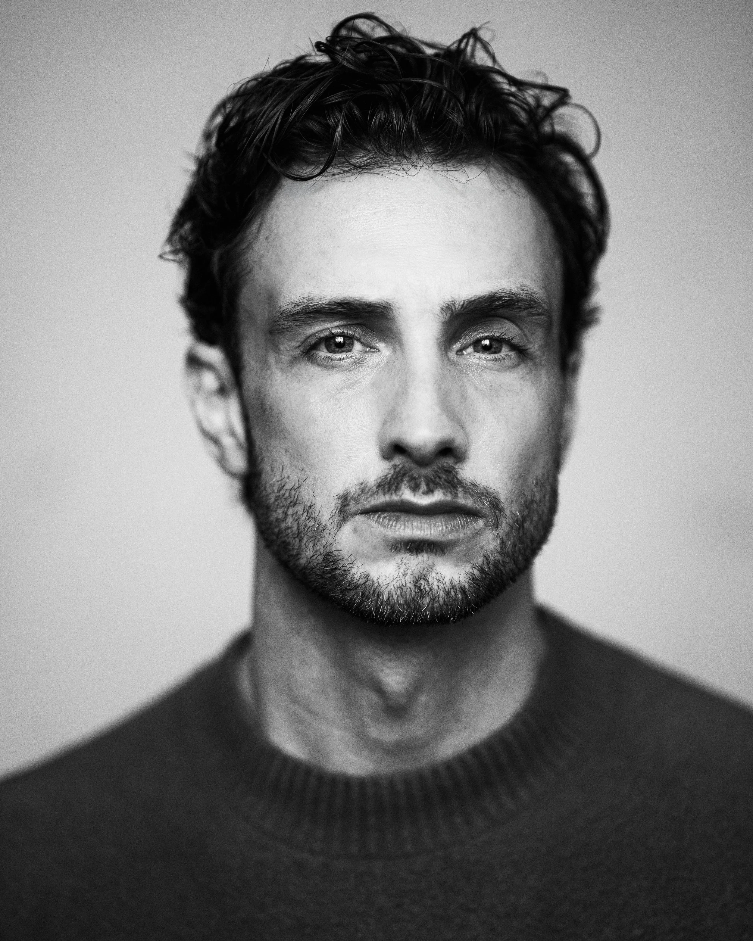Close-up portrait of a man with curly dark hair, blue eyes, a beard, wearing a white tank top and silver necklace, against a dark red background.