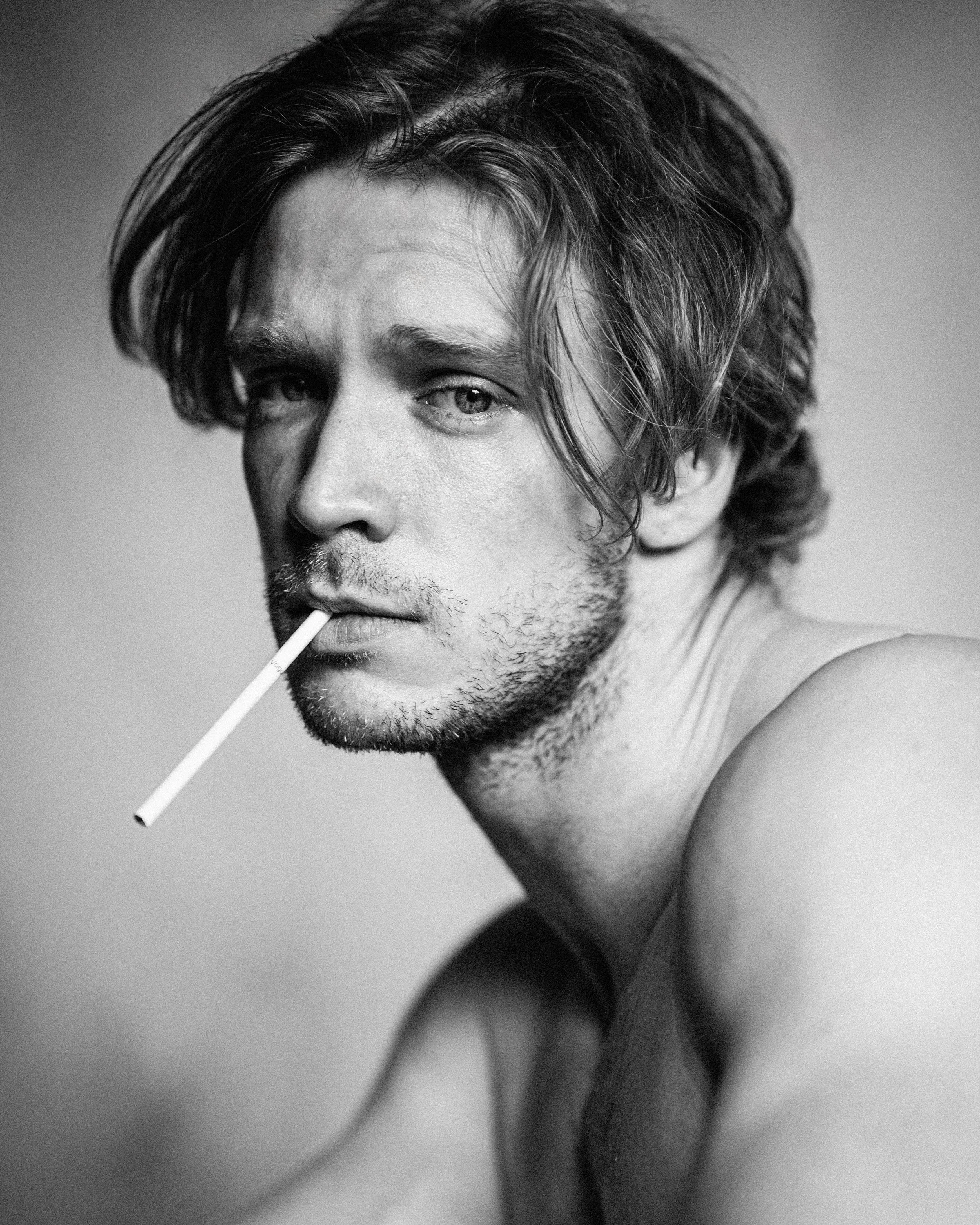 A black and white photo of a young man with messy hair, slight beard, and a cigarette in his mouth. He is looking at the camera with a serious expression, shirtless, with his arm visible in the foreground.
