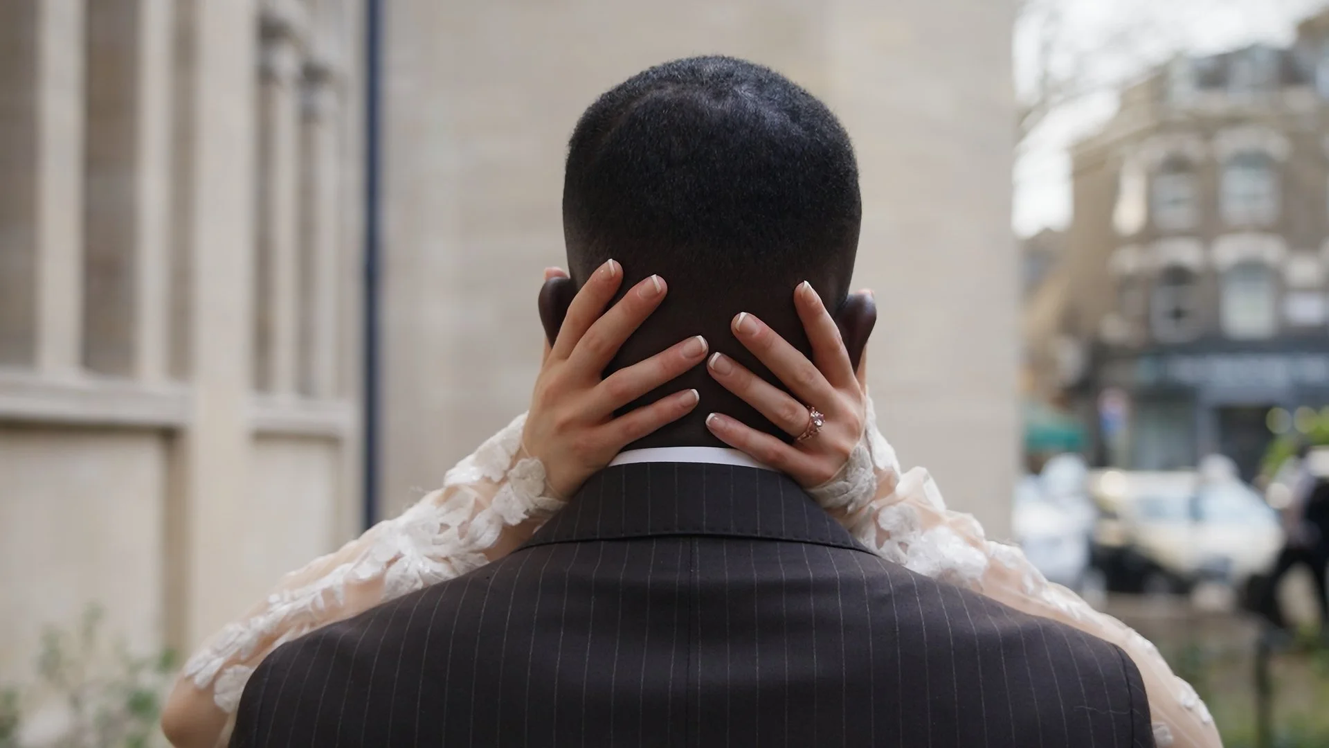 A couple embraces, with the woman's hands around the man's neck, seen from behind in an urban setting.