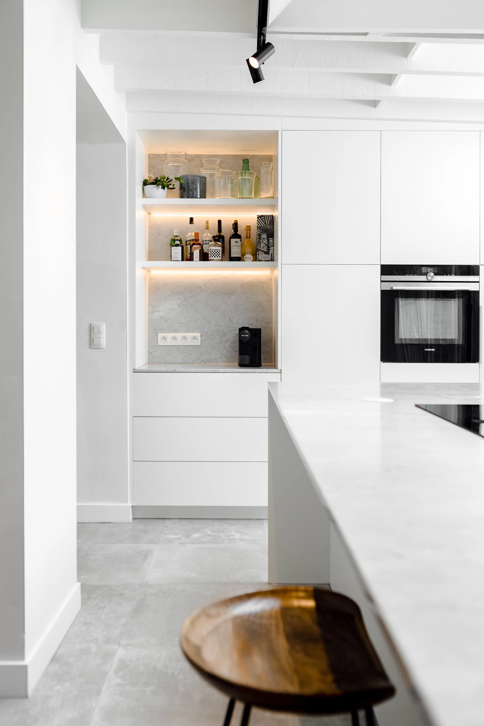 Moderne witte keuken met ingebouwde oven, open schappen met glazen en drankflessen, en een klein koffieapparaat. Het decor is minimalistisch en strak.