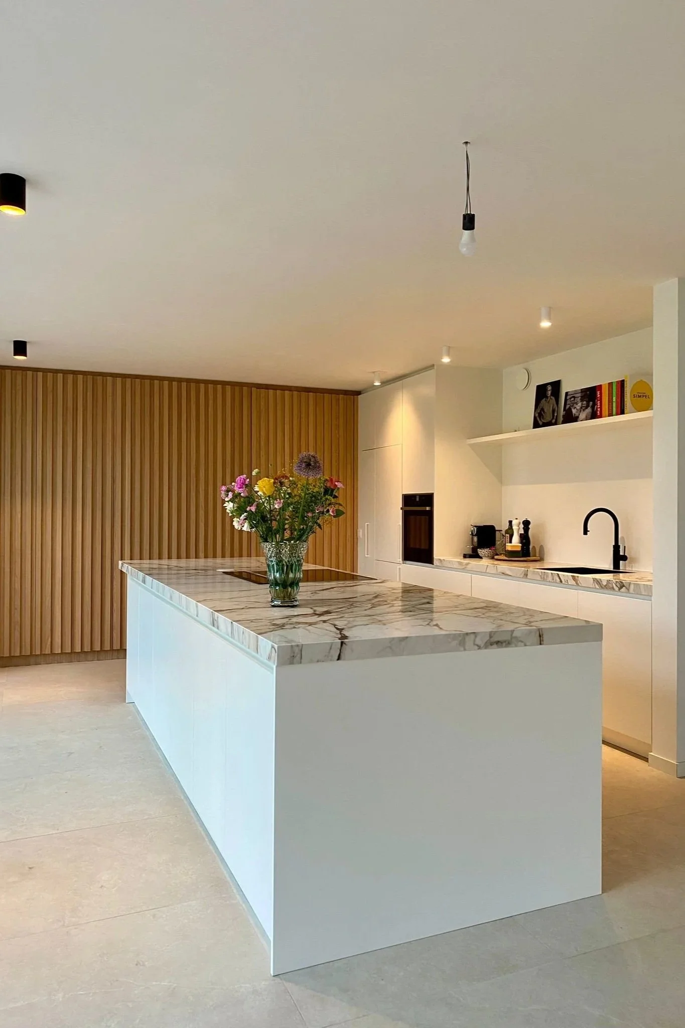 Moderne keuken met witte kasten, een marmeren keukenwerkblad en een centrale eiland met bloemen in een glazen vaas. In de achtergrond bevindt zich een houten verborgen kastenwand met ingewerkte pivot deur.