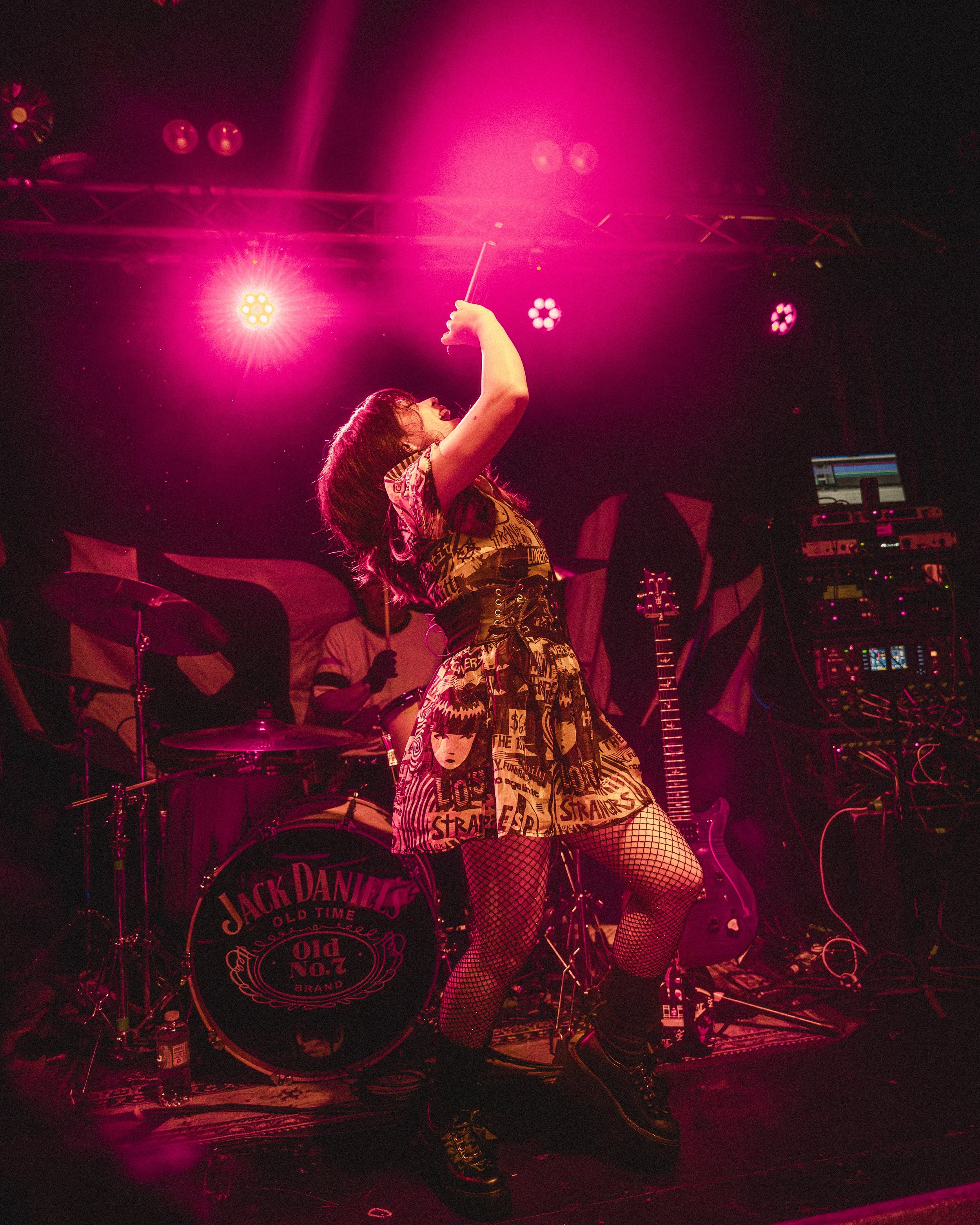 A woman singing and dancing on stage with pink lighting, drummer in background, guitar, and music equipment visible.