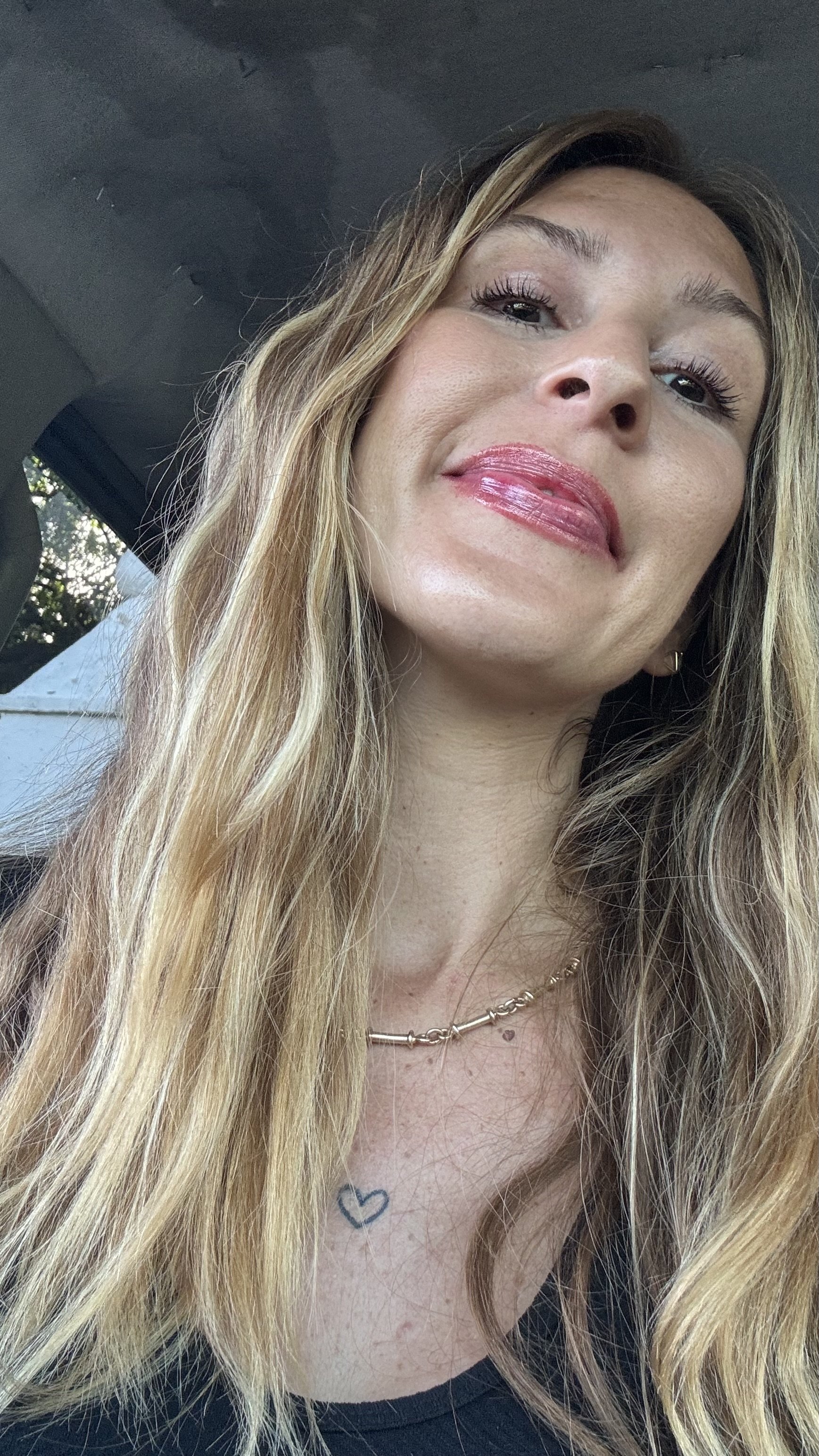 Close-up photo of a woman with long blonde hair, wearing pink lipstick, a necklace, and a black top. The photo is taken from a low angle, showing her face and part of her upper chest.