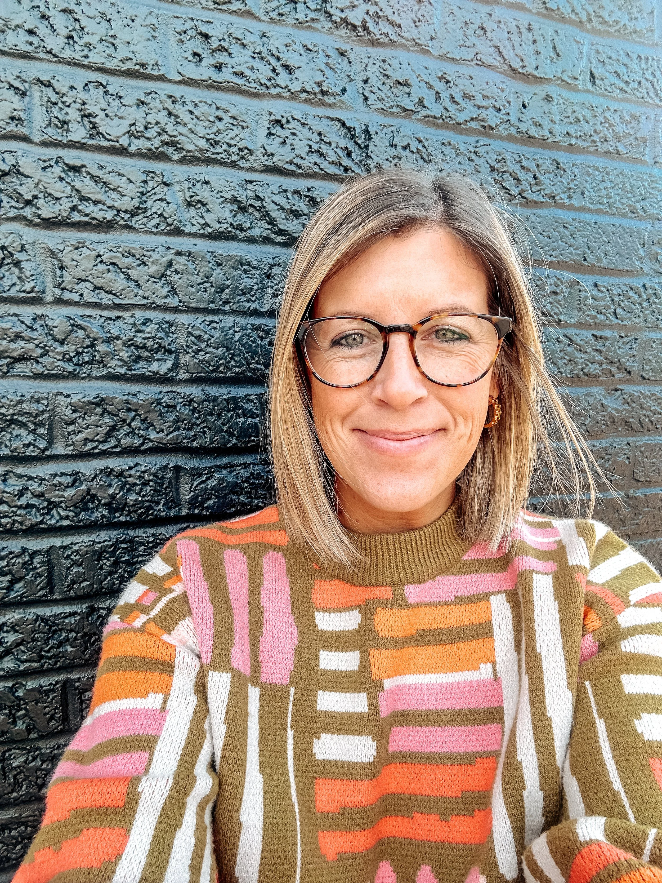 A woman with shoulder-length blonde hair wearing glasses and a colorful, patterned sweater. She is smiling and standing against a black brick wall.