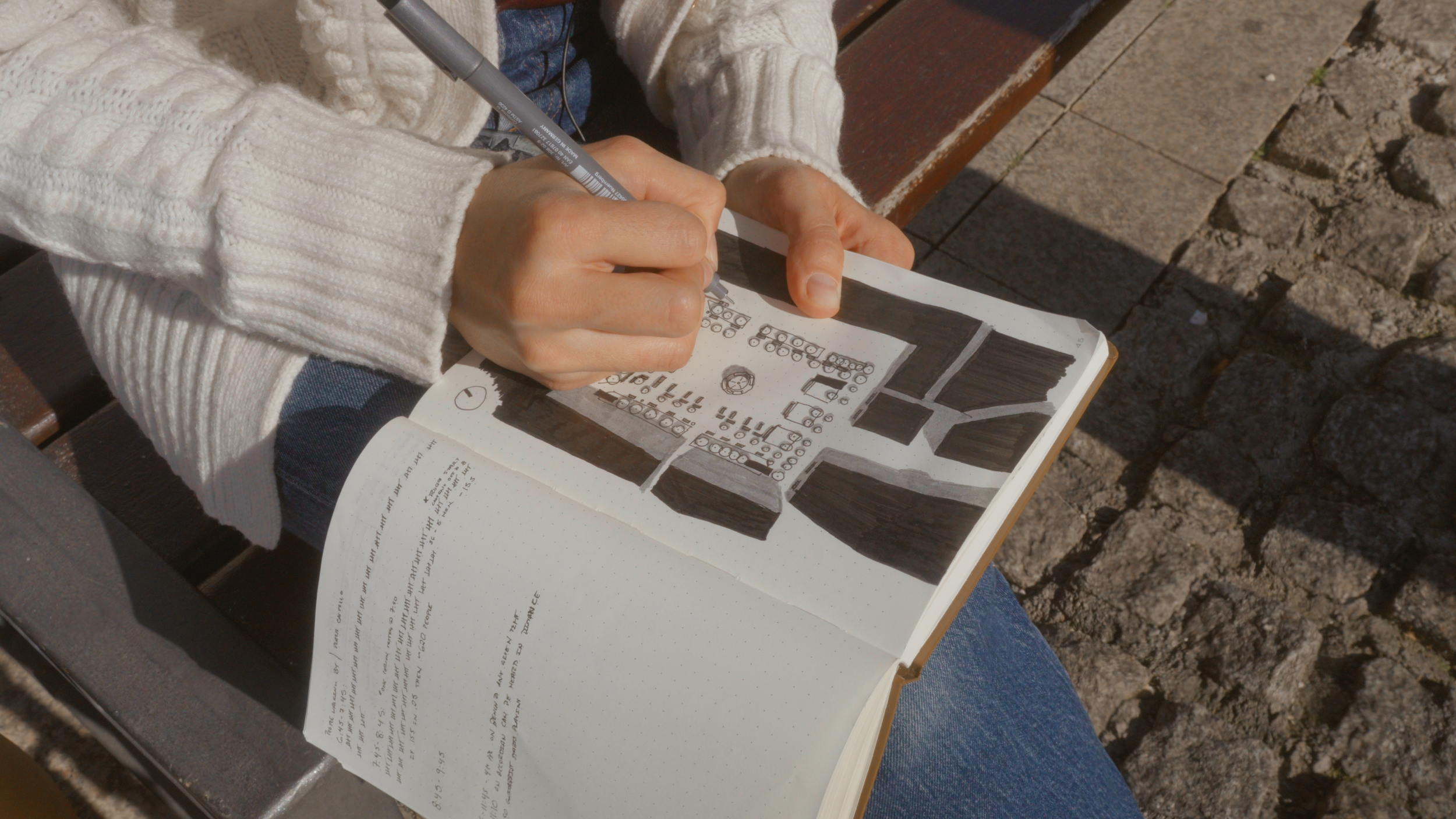 A person in a cream-colored sweater is sitting on a park bench, drawing or doodling in a sketchbook with black ink, with a paved stone walkway visible beside them.