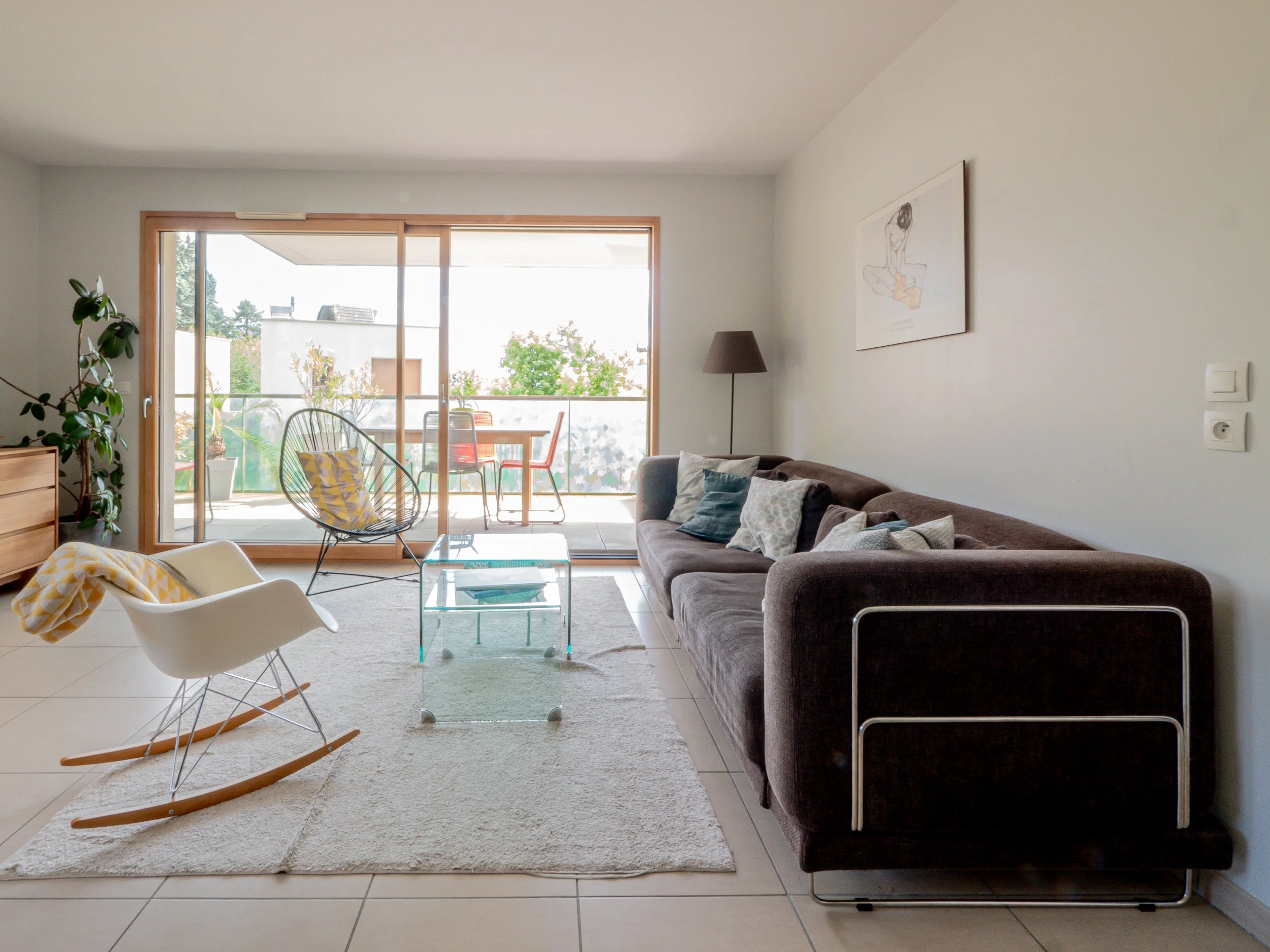 Salon moderne avec canapé marron, rocking chair blanc, tapis beige, table en verre, vue sur balcon avec mobilier extérieur, plantes vertes, tableau abstrait au mur, lampe de sol