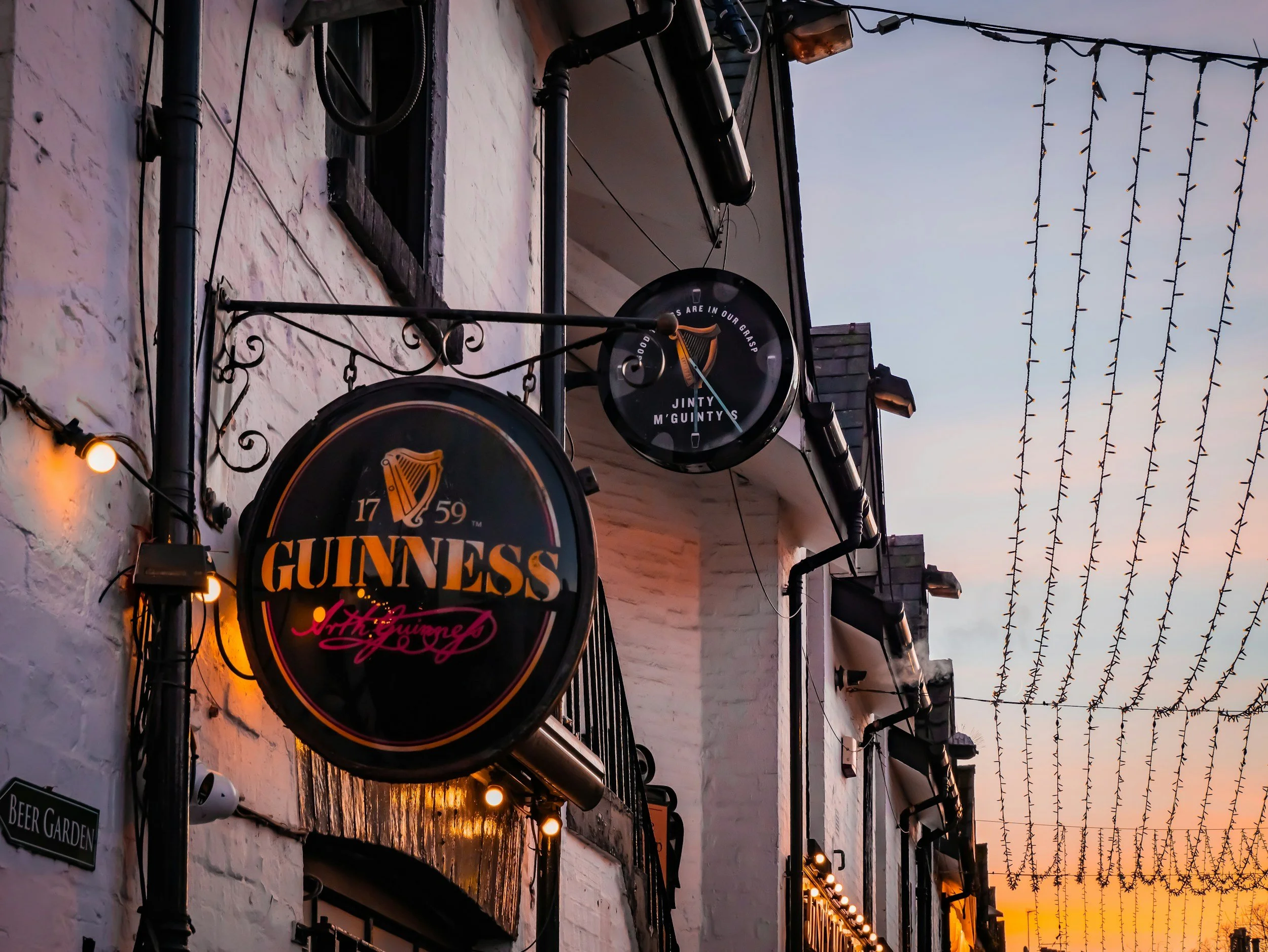 Exterior view of a pub or bar with Guinness signage, decorative hanging lights, a white brick wall, and a sunset sky with string lights overhead.