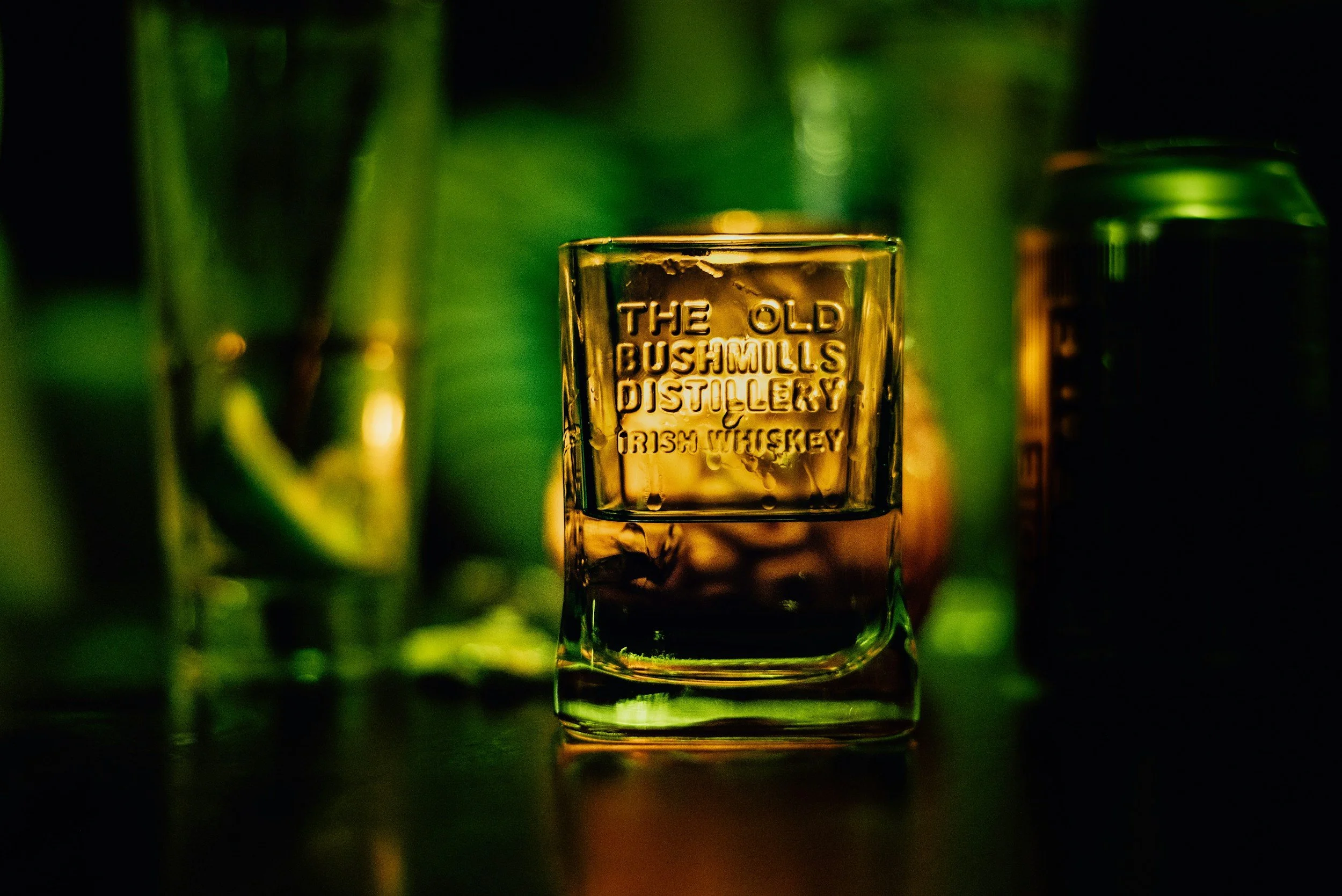A glass of whiskey with embossed text, surrounded by green bottles and a soda can, in a dimly lit setting.