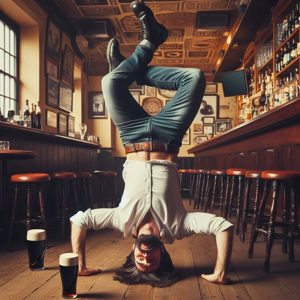 A man with long hair and a beard performing a handstand on the wooden floor of a bar or pub, with two glasses of dark beer nearby. He is wearing a light-colored shirt, jeans, and black shoes, and is upside down in the center of the image.