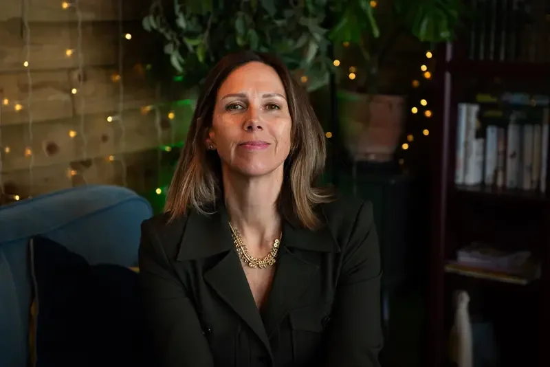 A woman with shoulder-length brown hair sitting on a blue couch in a cozy, warmly lit room with fairy lights, a wooden wall, and a bookshelf in the background.