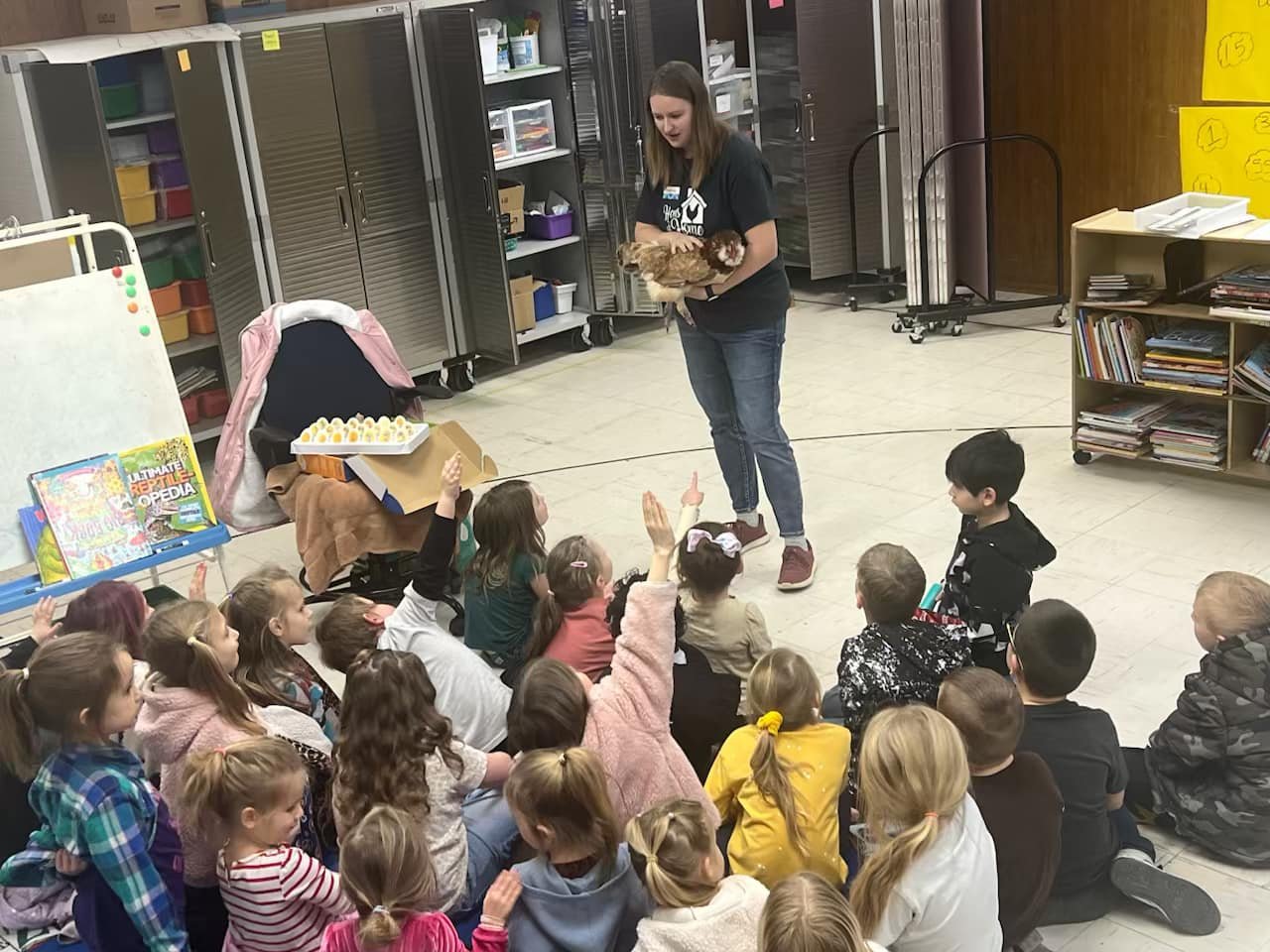 A woman holding a chicken in front of a group of children sitting on the floor, with some children raising their hands in a classroom setting.