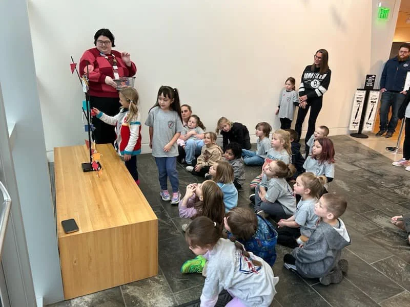 A group of children seated on the floor and some standing, watching a woman demonstrate a toy on a wooden table, with adult onlookers in the background.