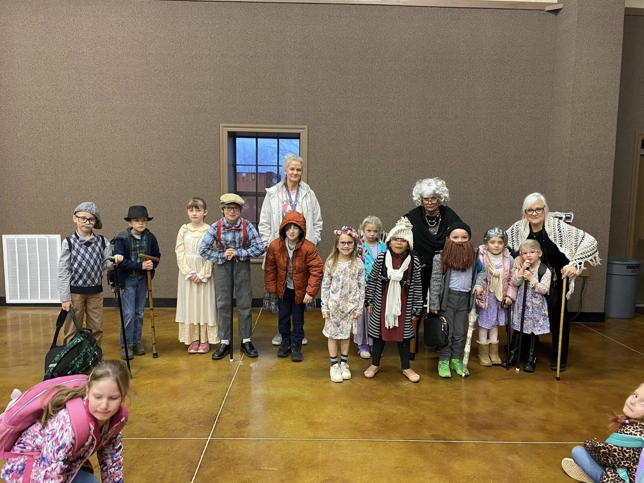 Group of children dressed in costumes, standing with two women in costume, in a room with a brown wall, wood floor, and a window in the background.