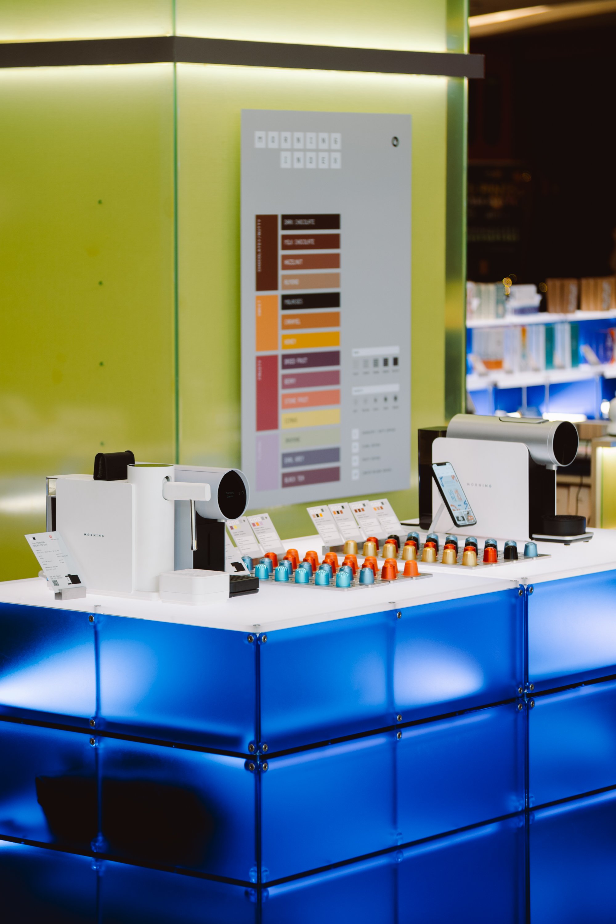 Coffee capsule vending machine with various coffee pods displayed on a white counter, with a smartphone on a stand, in a modern bright space.