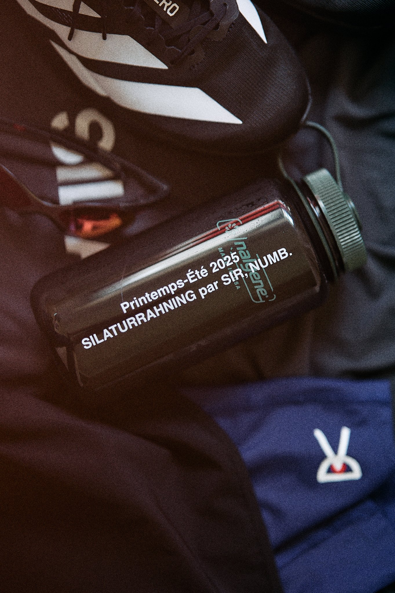 A black water bottle with white and teal text resting on dark clothing. The text on the bottle reads: 'Printemps-Été 2025. SILATURRAHNing par SIR, NUMB.' The surrounding clothing includes a pair of black sneakers with white accents, a pair of sunglas