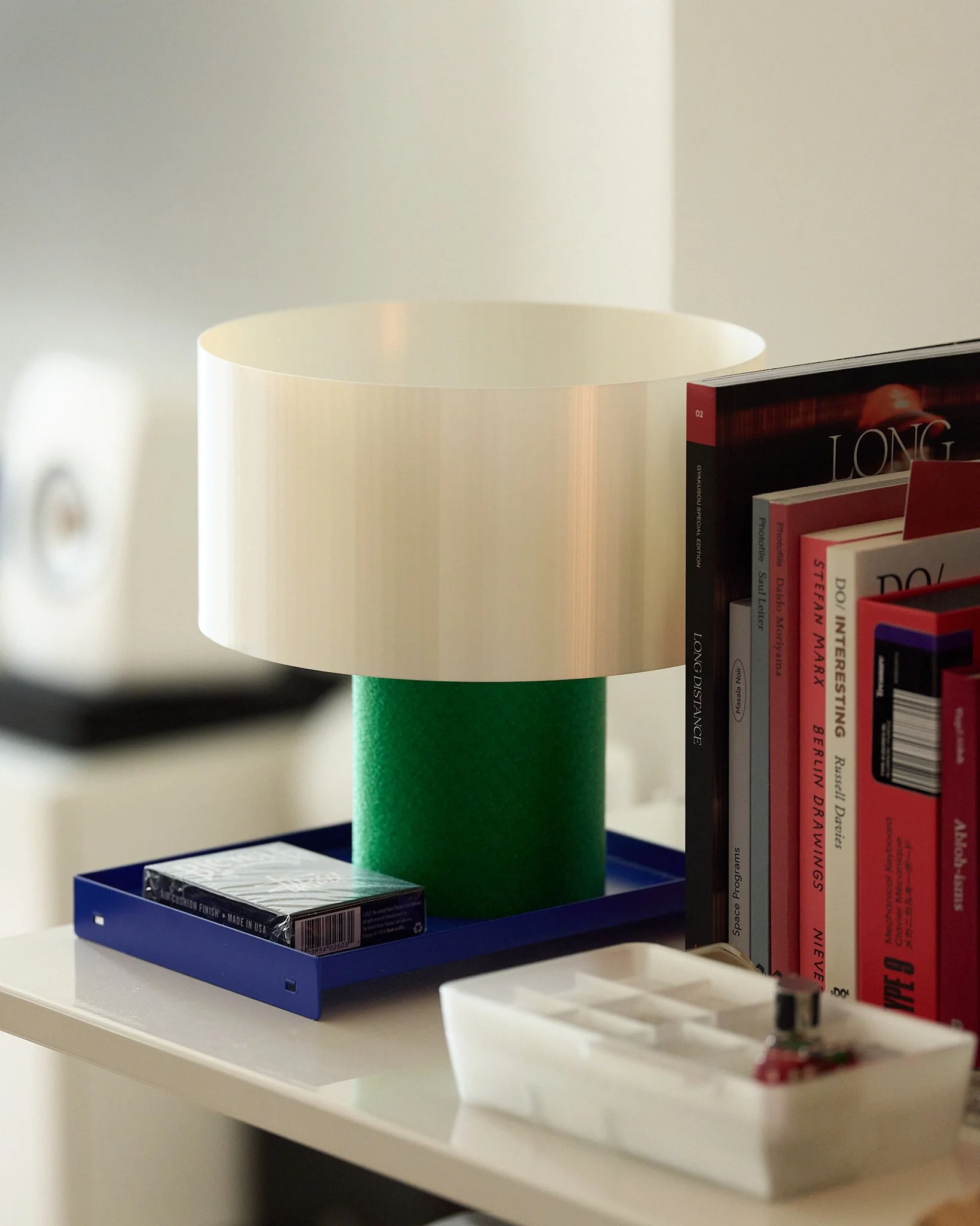 A modern table lamp with a white cylindrical shade, a green column base, placed on a white surface next to a bookshelf with various books and a small dish.