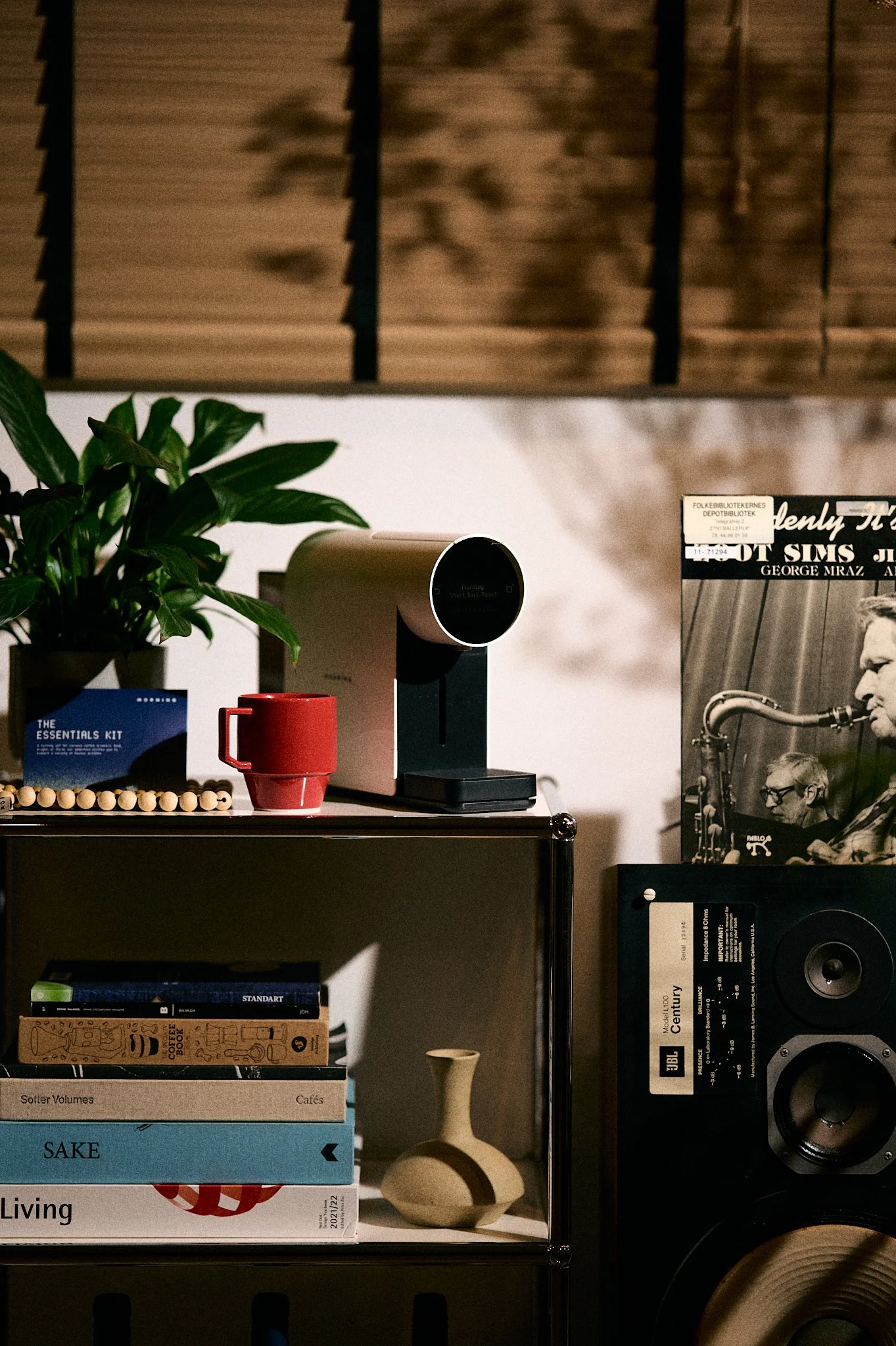 A shelf displaying a potted plant, a red mug, a small beige decorative vase, and stacked books with a speaker on the right side, with a wooden design overhead.
