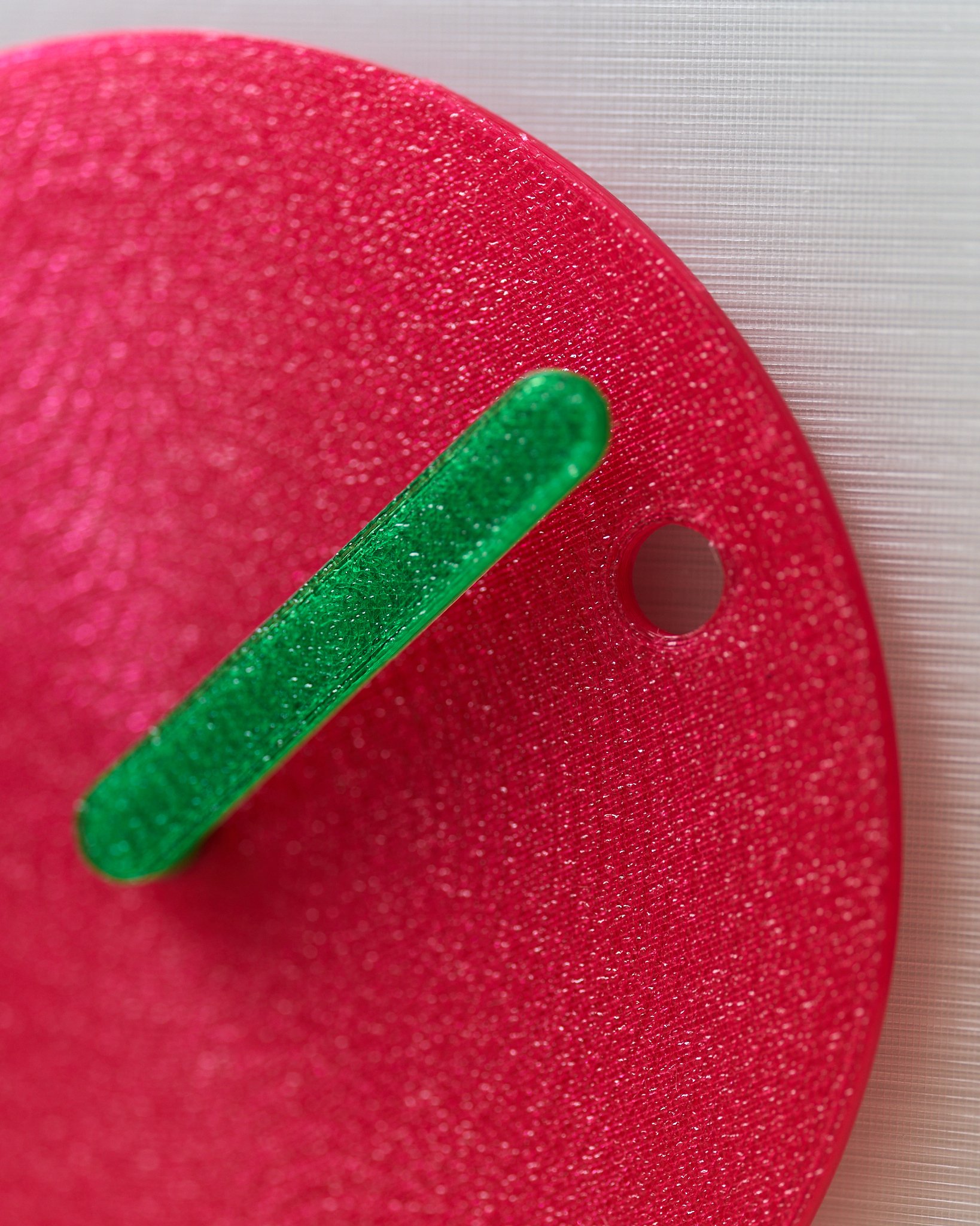 Close-up of a pink, glittery wall clock with a green clock hand, mounted on a light-colored textured wall.