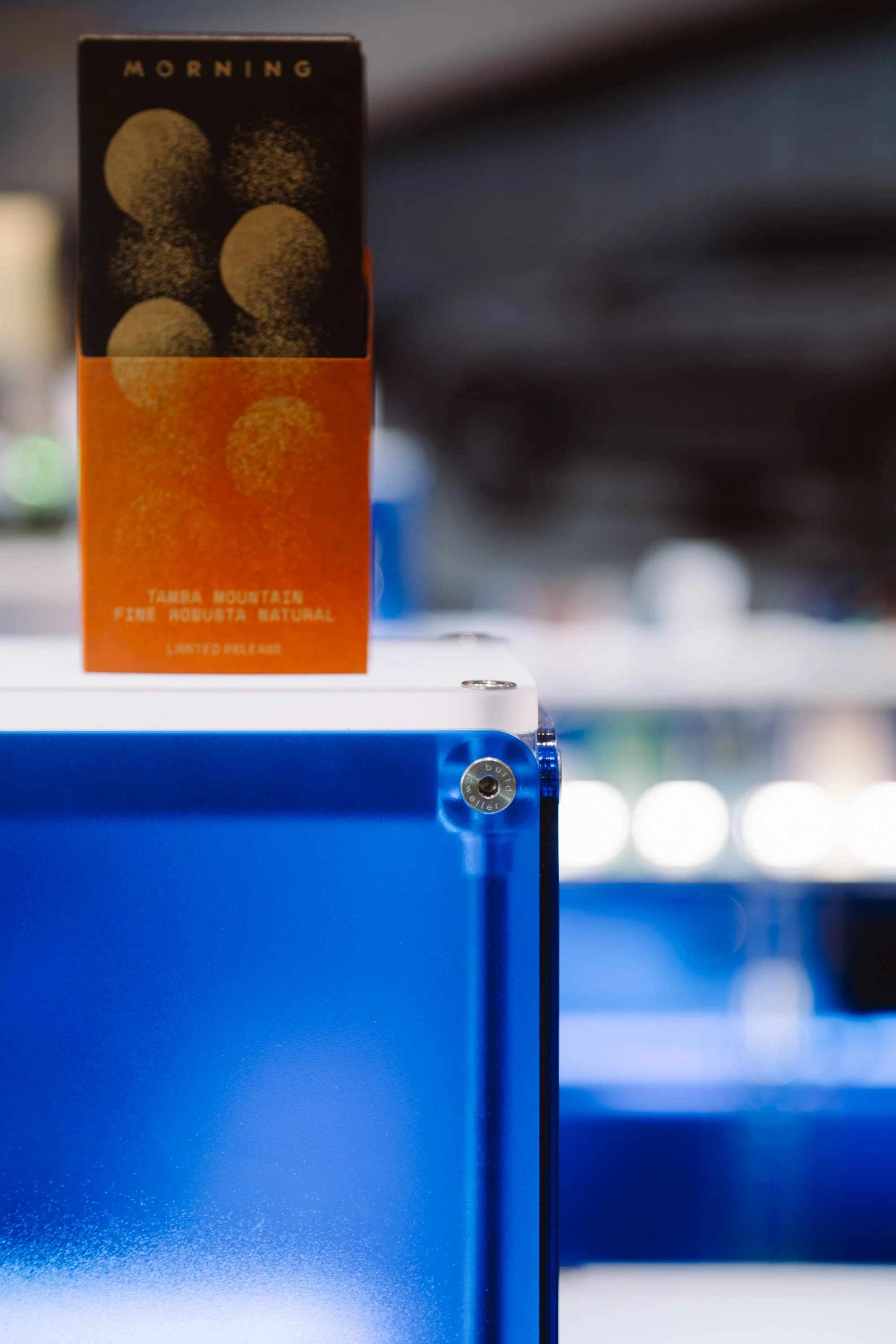 A carton of Maui Mountain coffee placed on a blue refrigerator in a store or showroom. The carton features a black top section with images of coffee beans and the word 'Morning,' and an orange bottom section with text indicating it is a limited relea