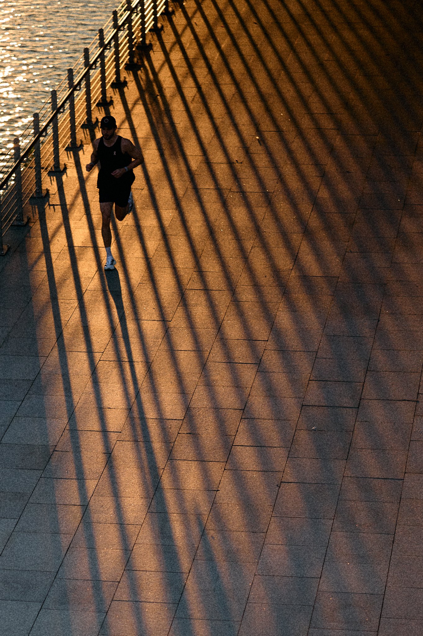 A person jogging along a pathway near a body of water, with long shadows cast by a fence or railing during sunset.