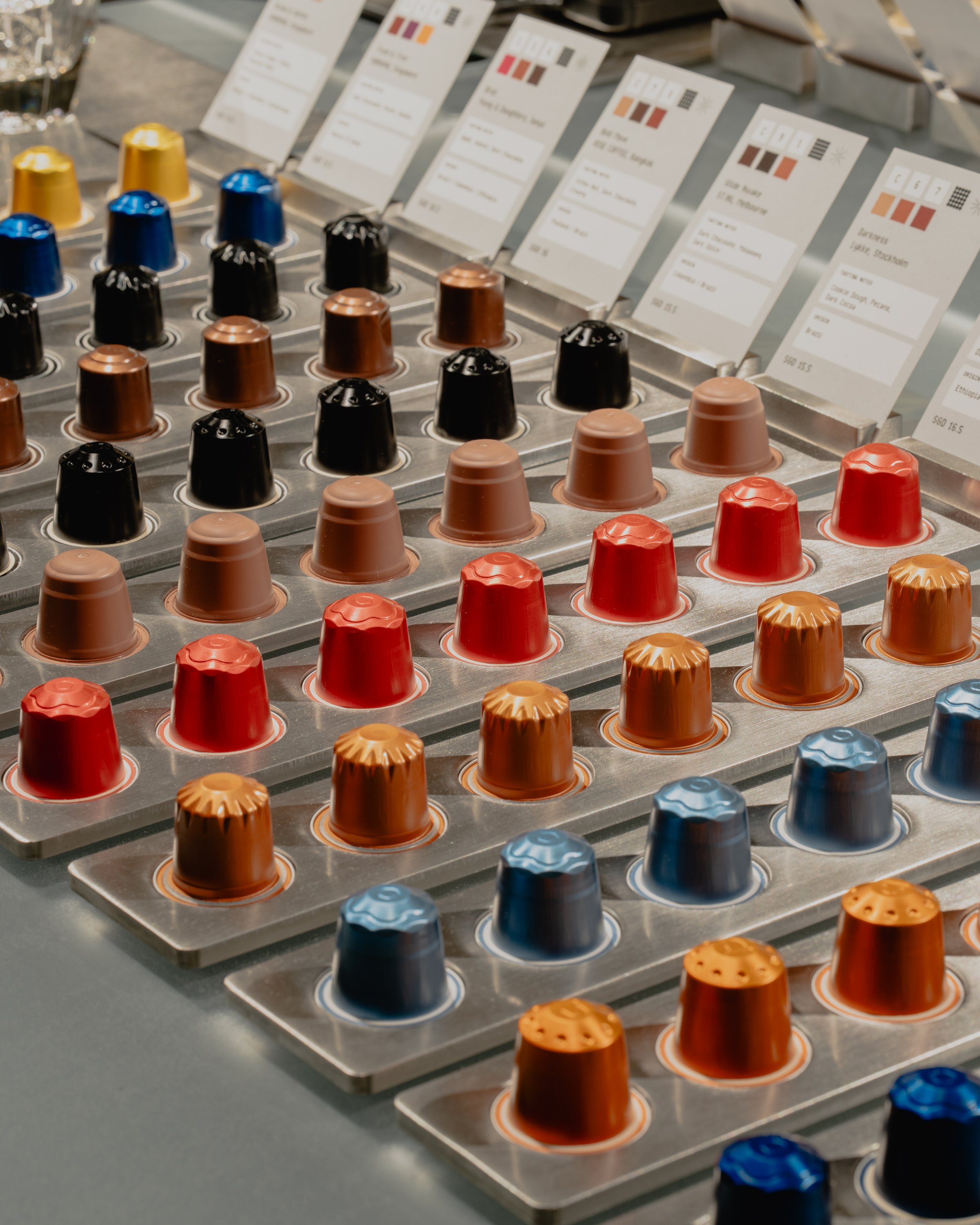 A display of various coffee capsules arranged in rows on a metallic tray with informational cards in the background.