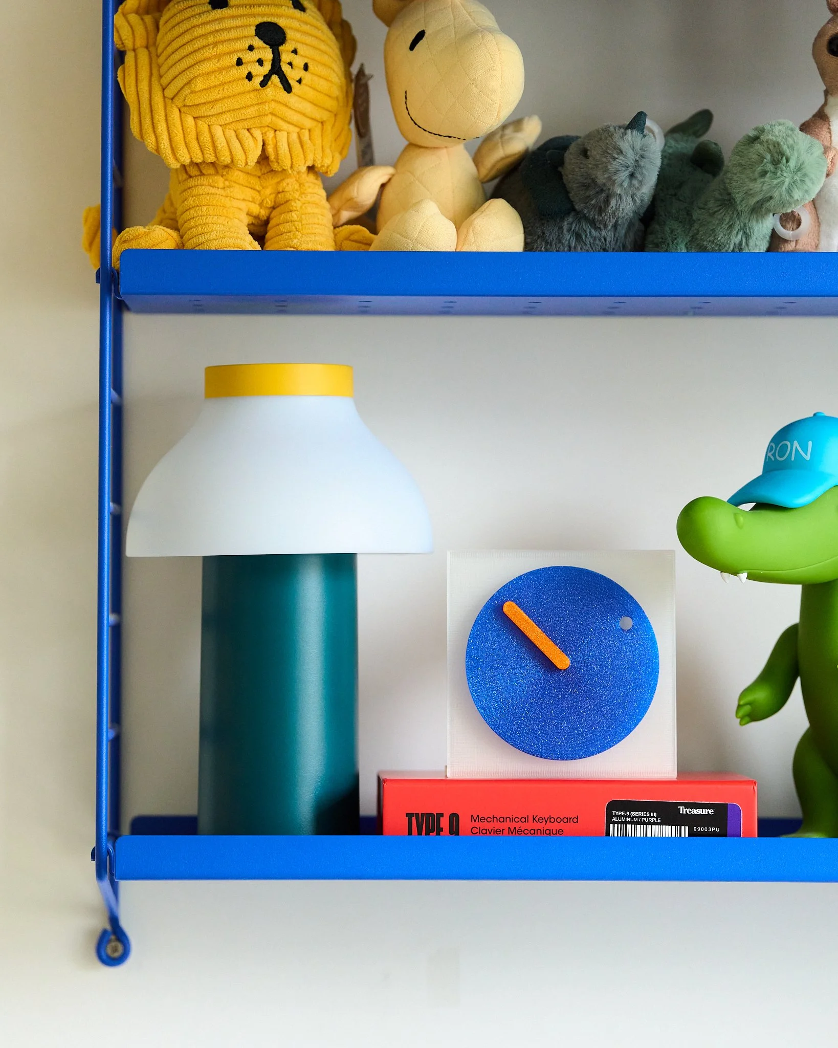 Blue wall shelf with plush toys resembling lions and dinosaurs on top, a modern lamp, a blue clock with an orange hand, and a red box labeled 'Type Q' underneath.