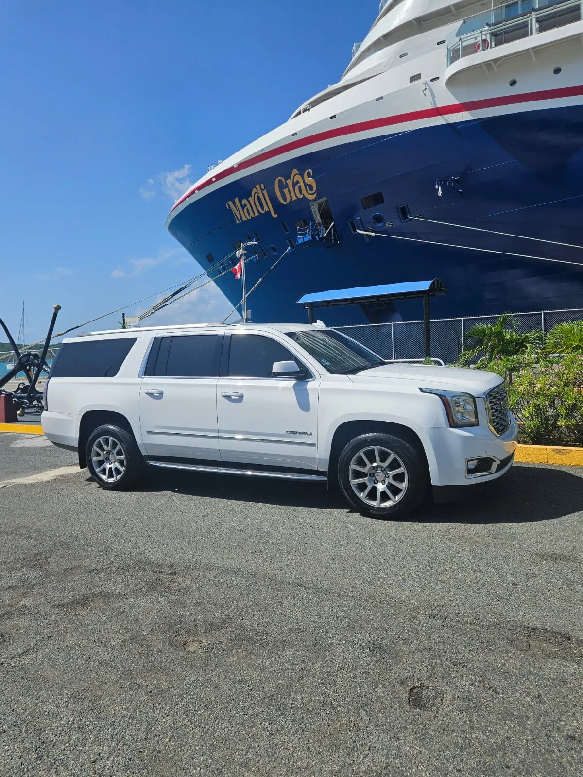 A white SUV parked on a paved area near a large blue and white cruise ship named Mardi Gras, with a clear blue sky in the background.