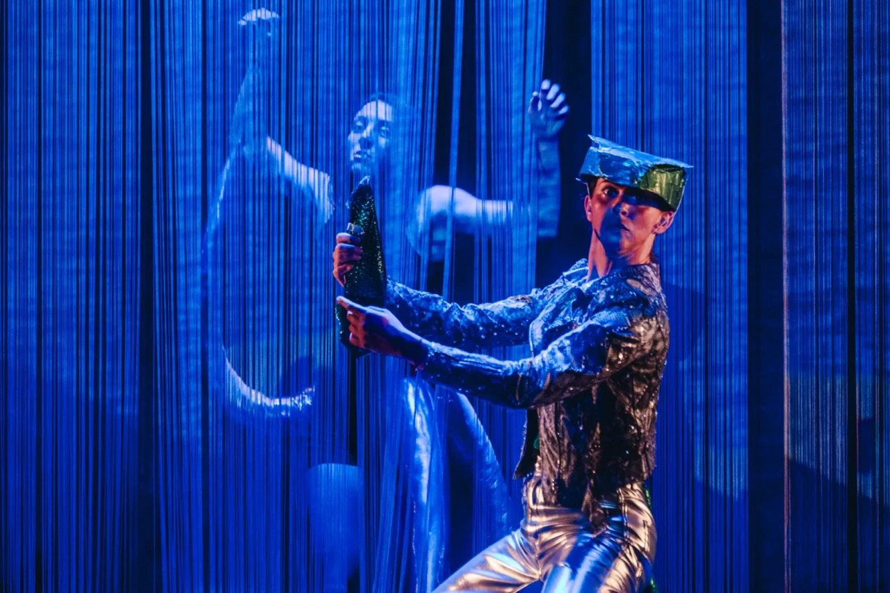 Deux performeurs sur scène dans un décor futuriste, avec des vêtements métallisés, entourés de rideaux fins et lumière bleue.