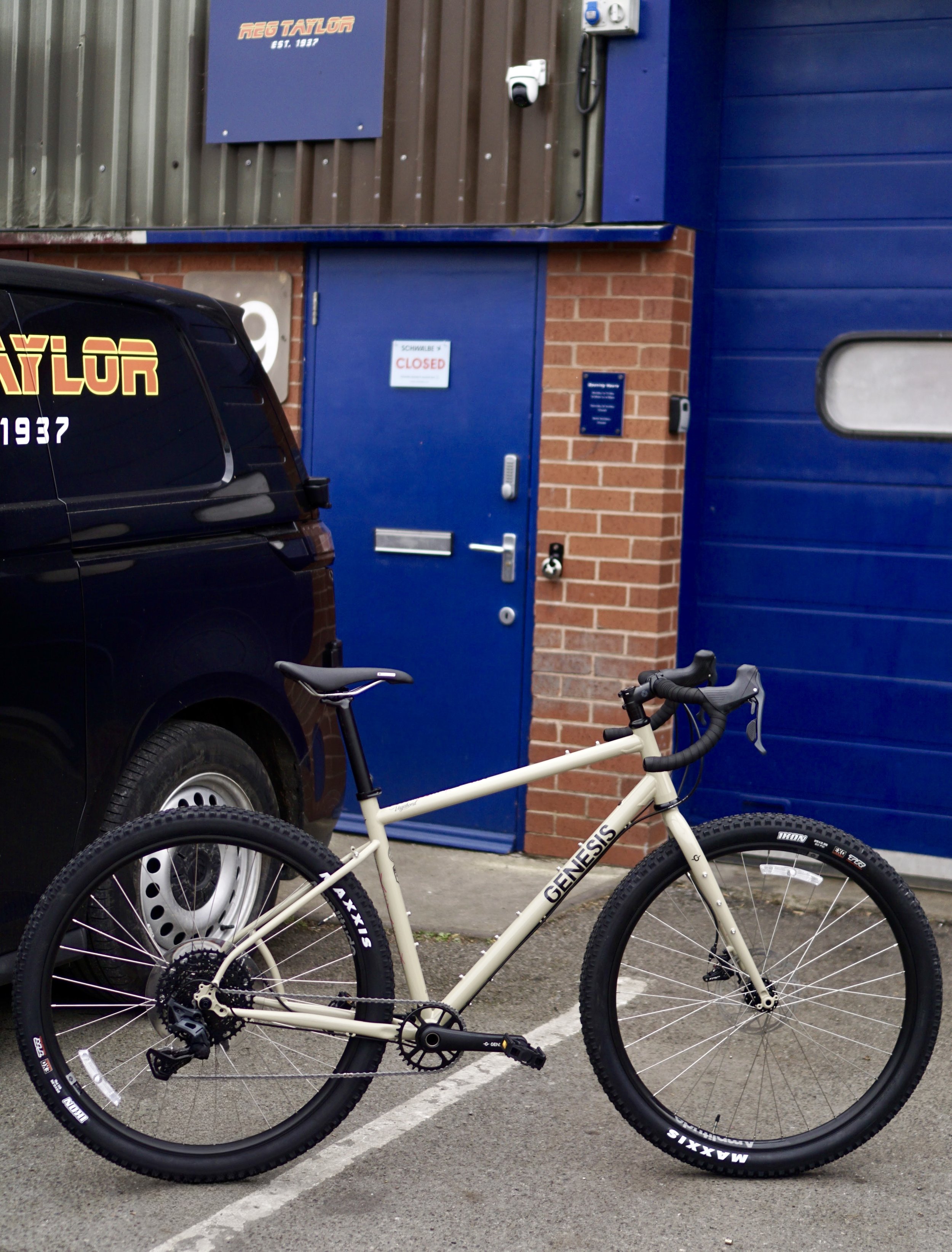 A white Genesis mountain bike with black tires and a black saddle is parked against a yellow painted parking lot line near a blue garage door and a black van. The background shows a brick wall and a sign that reads "NEO TAYLOR" with a smaller sign in