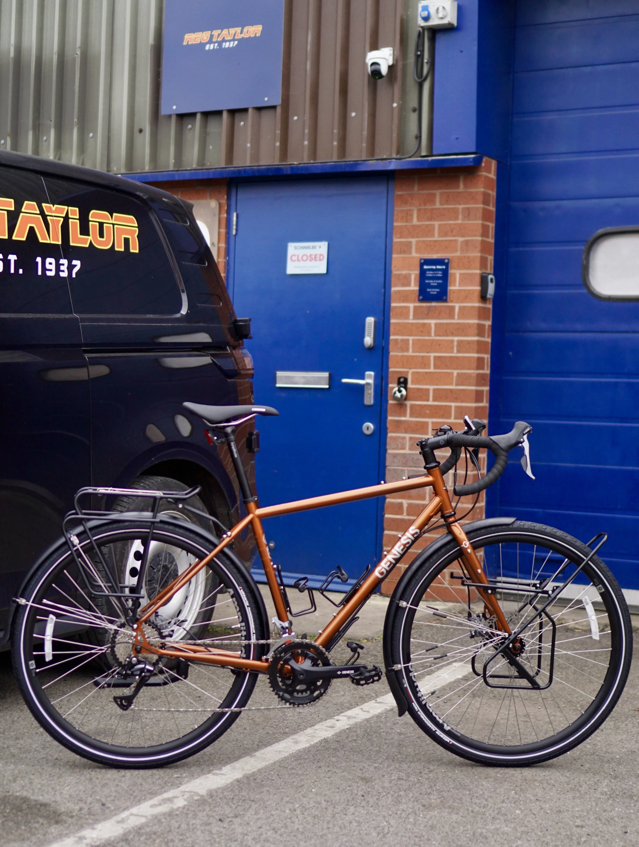 A brown Genesis hybrid bicycle with front and rear racks is parked in a parking lot in front of a blue door with a sign that reads 'Closed.' A black van is parked next to the bike, partly blocking the view of the building behind it, which has a sign 