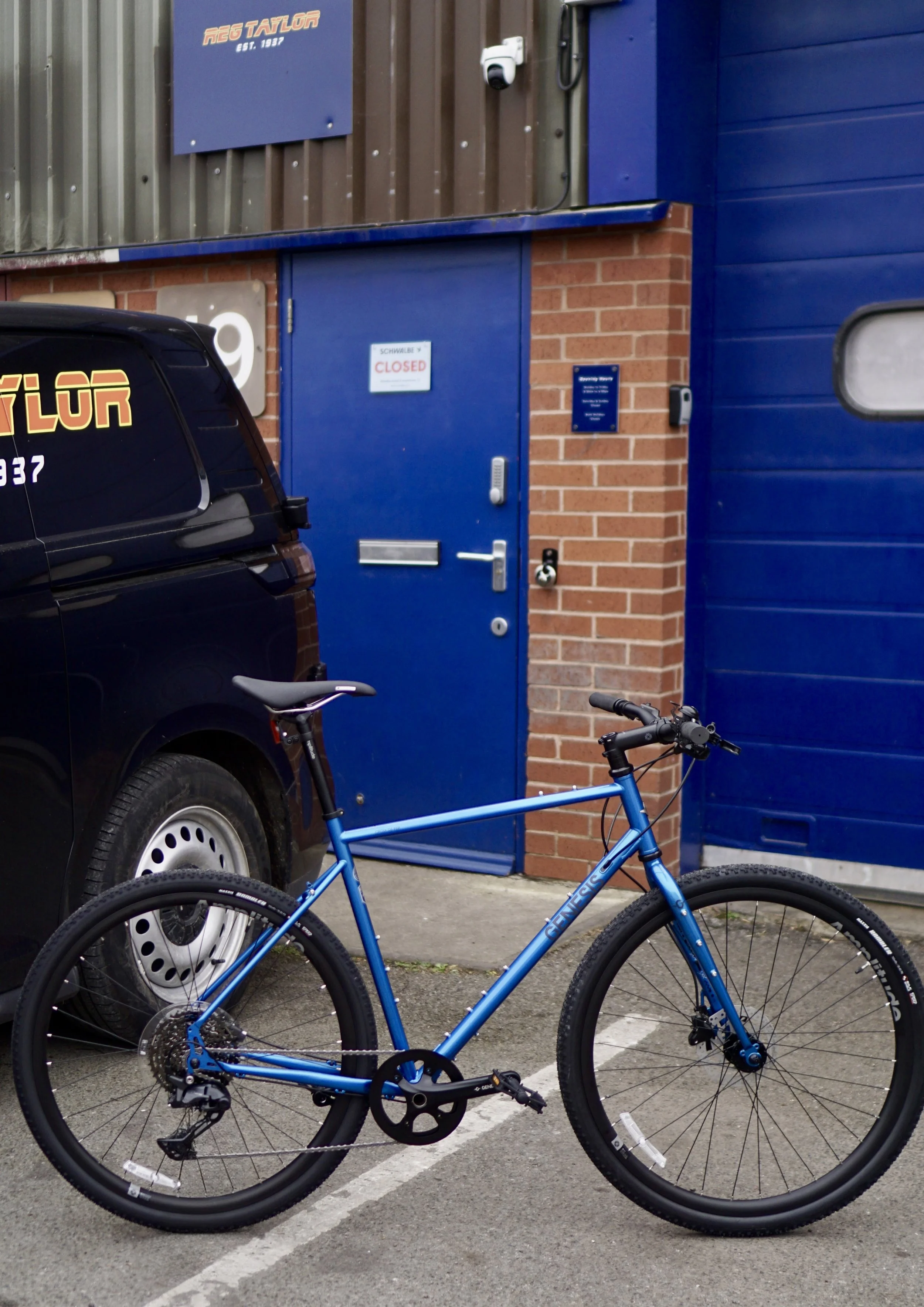 A blue bicycle parked in a parking lot next to a black van, against a background of a building with a blue door, a brick wall, and a sign indicating "CLOSED."