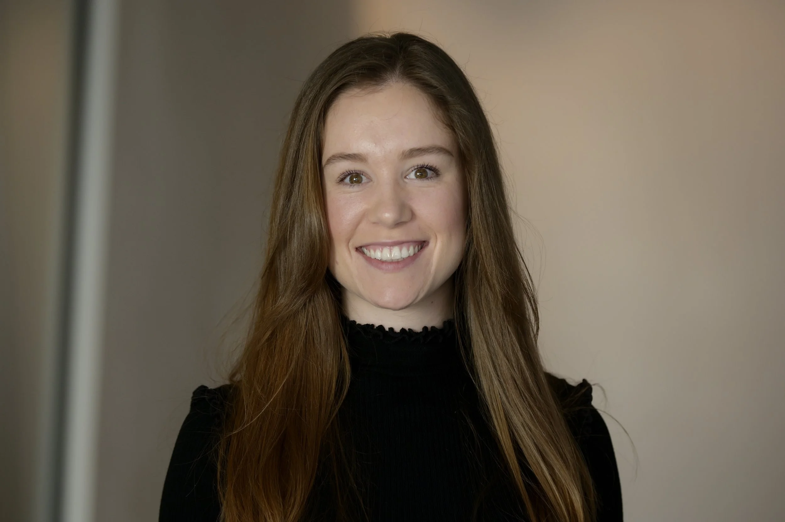 A smiling man with short light brown hair and blue eyes, wearing a navy blue blazer over a white shirt, standing against a dark textured wall.